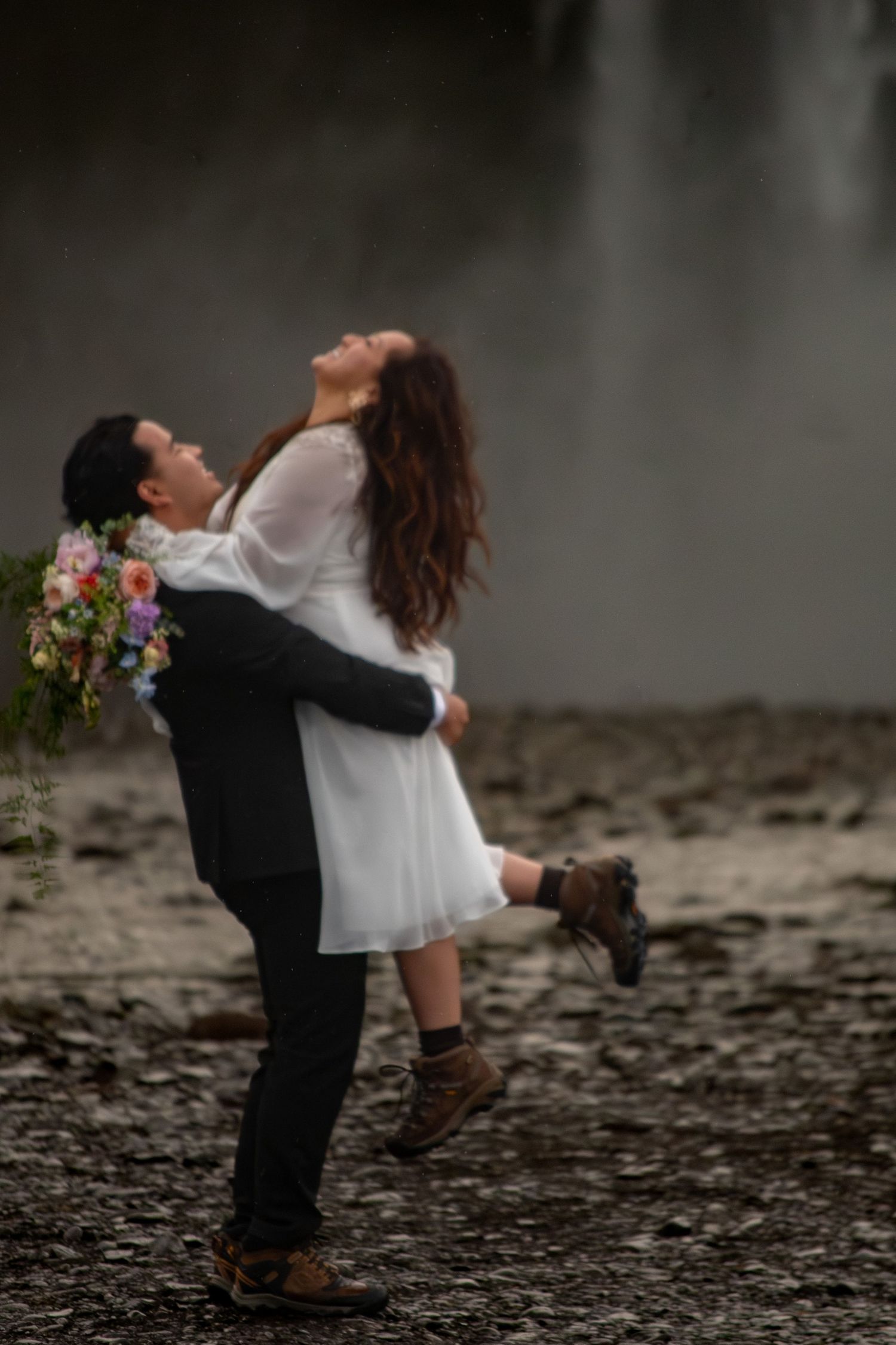 Groom lifting bride in white dress outdoors near waterfall, she holds colorful bouquet, both wearing hiking boots.