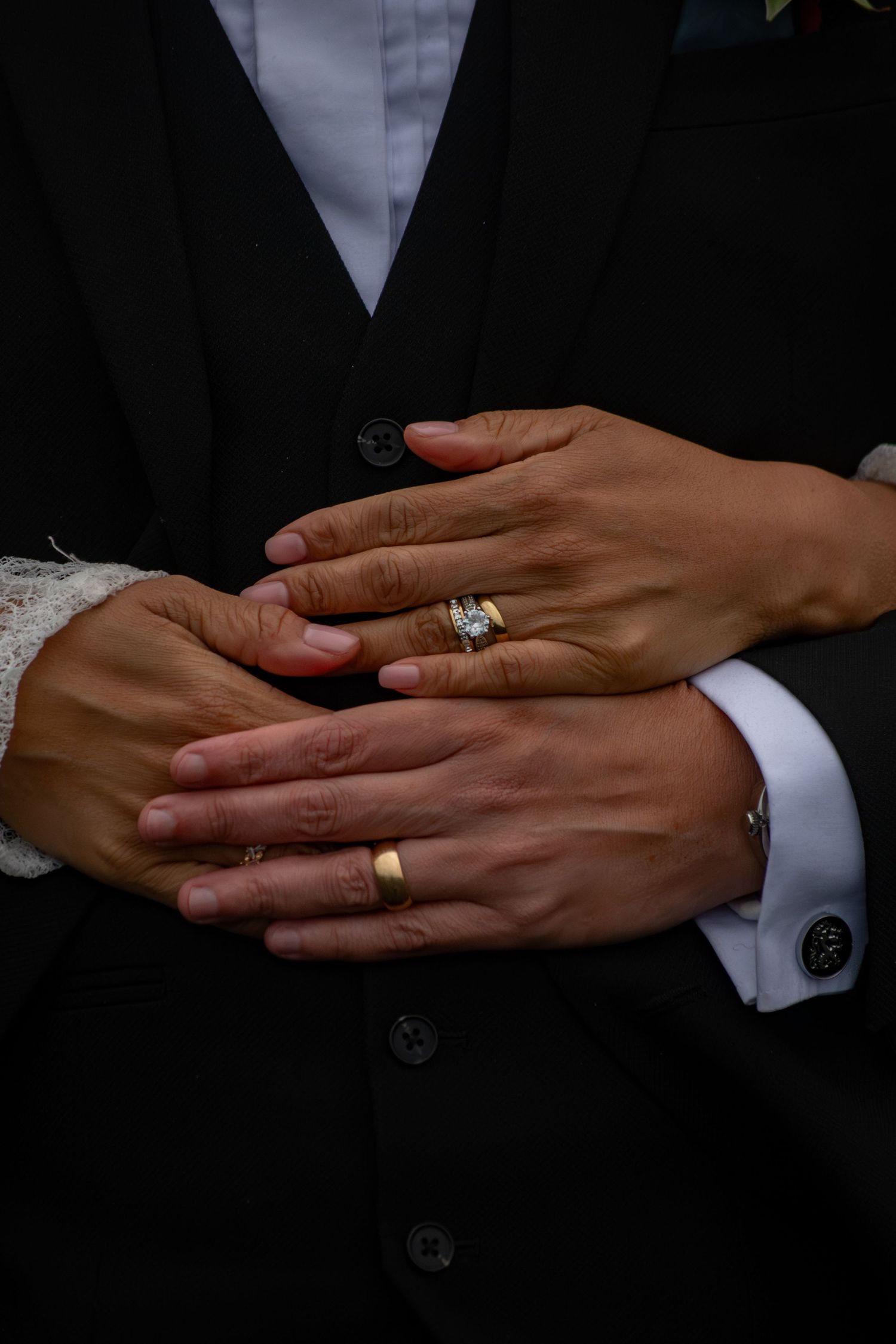Close-up of newlywed couple's hands showing gold wedding bands and diamond engagement ring against black suit.