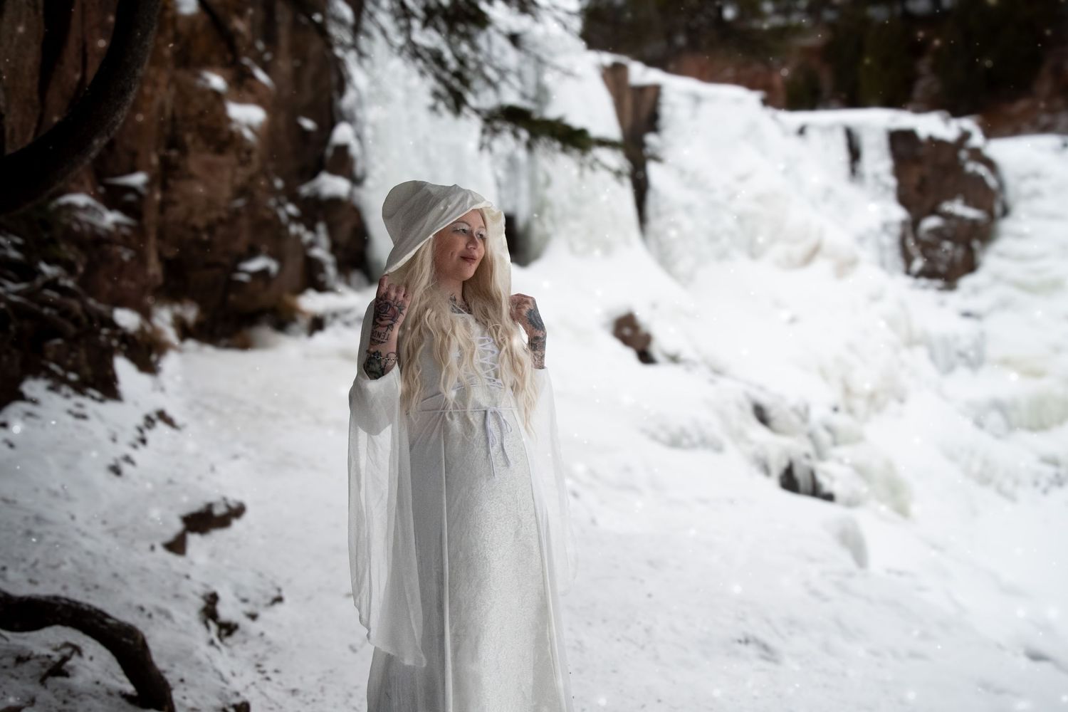 Blonde woman in white hooded cape dress praying near frozen waterfall in winter.