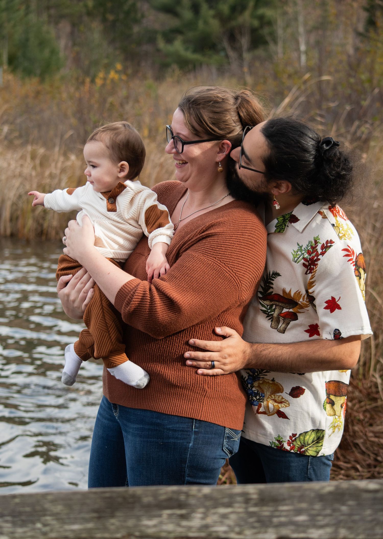 A happy young family with a mother holding a baby while a father kisses her cheek outdoors near a lake.