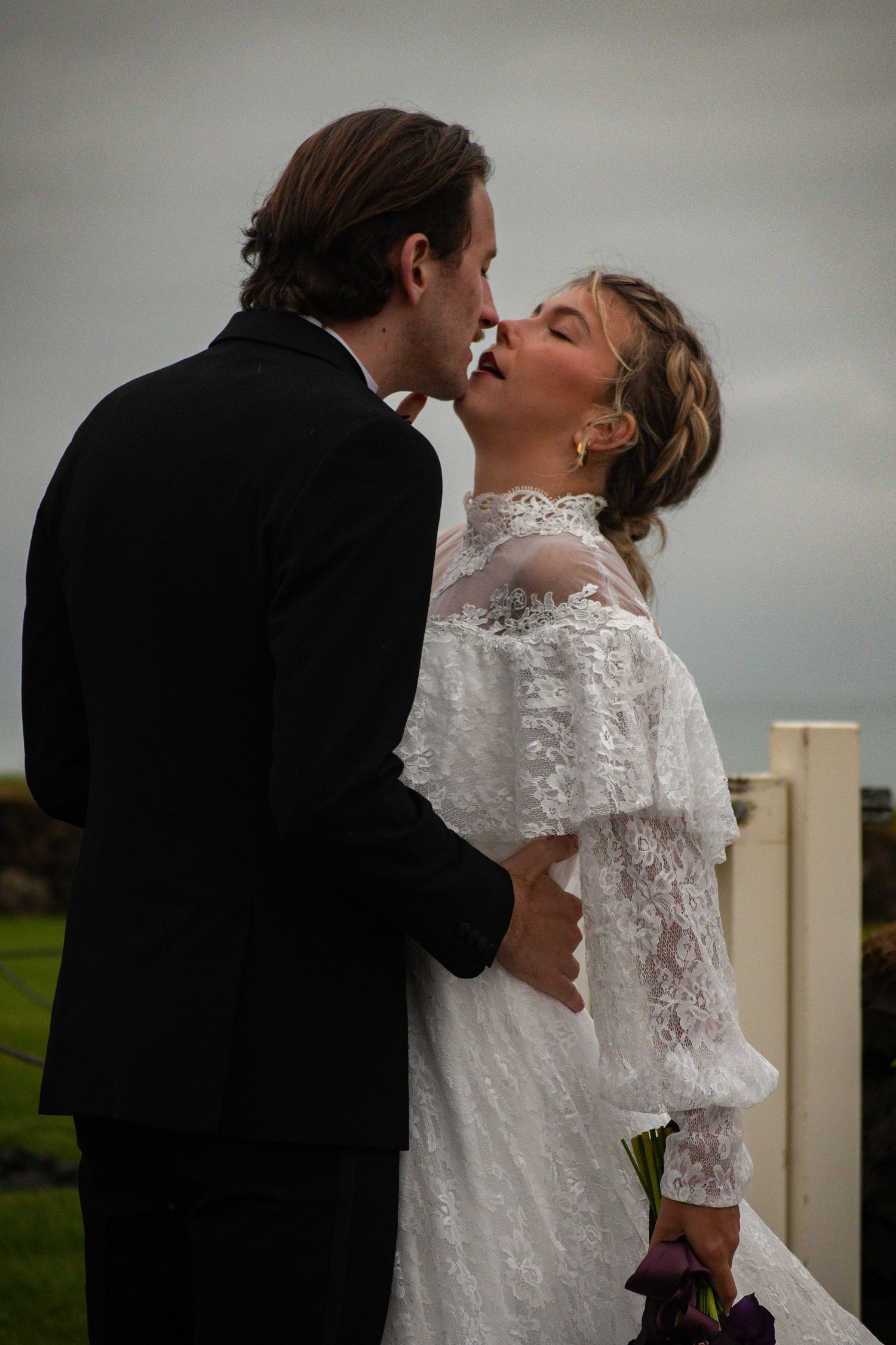 A bride and groom share a romantic kiss outdoors, she in lace dress holding flowers, he in black suit, on their wedding day.