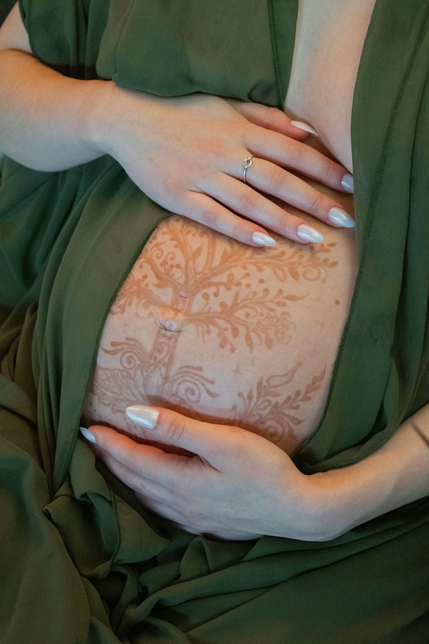 Pregnant woman in green dress with henna belly art, hands cradling bump, wearing engagement ring with white manicure.