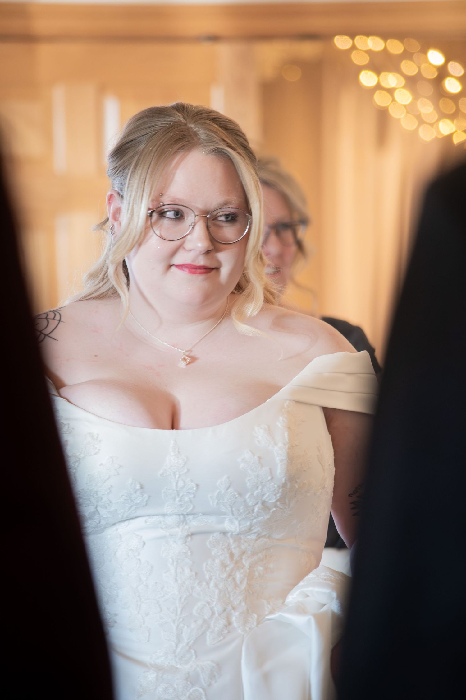 Blonde bride in off-shoulder lace wedding gown wearing glasses smiles warmly during wedding preparations.