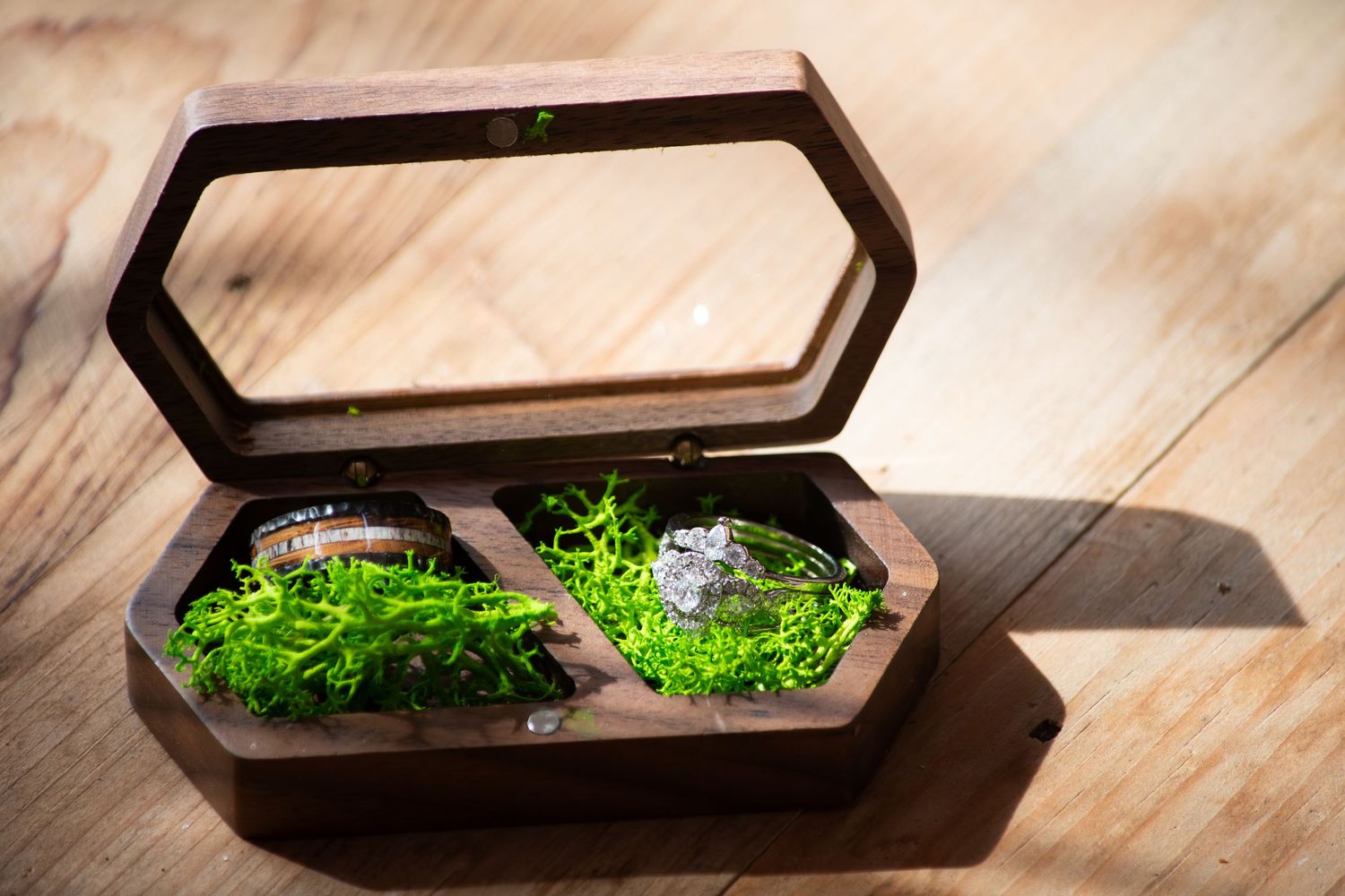 Two wedding rings displayed in a rustic wooden box with green moss on a wooden surface.