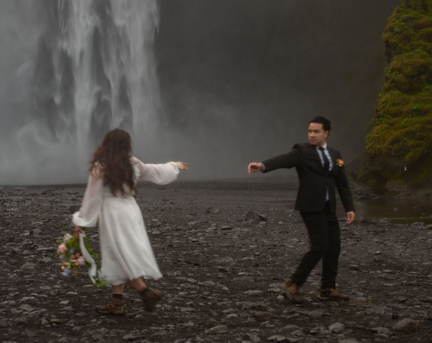 Bride and groom dancing near a majestic waterfall in Iceland, wearing wedding attire on a rocky shore.
