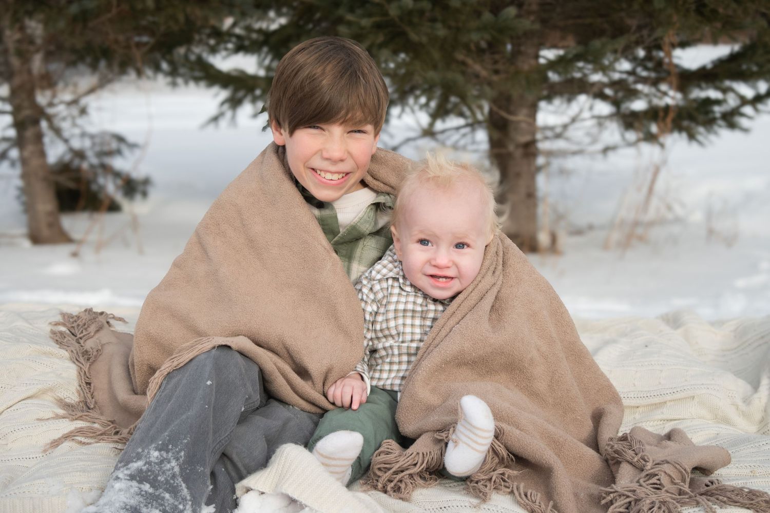 Two siblings wrapped in a tan blanket sitting in snow, older boy hugging smiling baby.