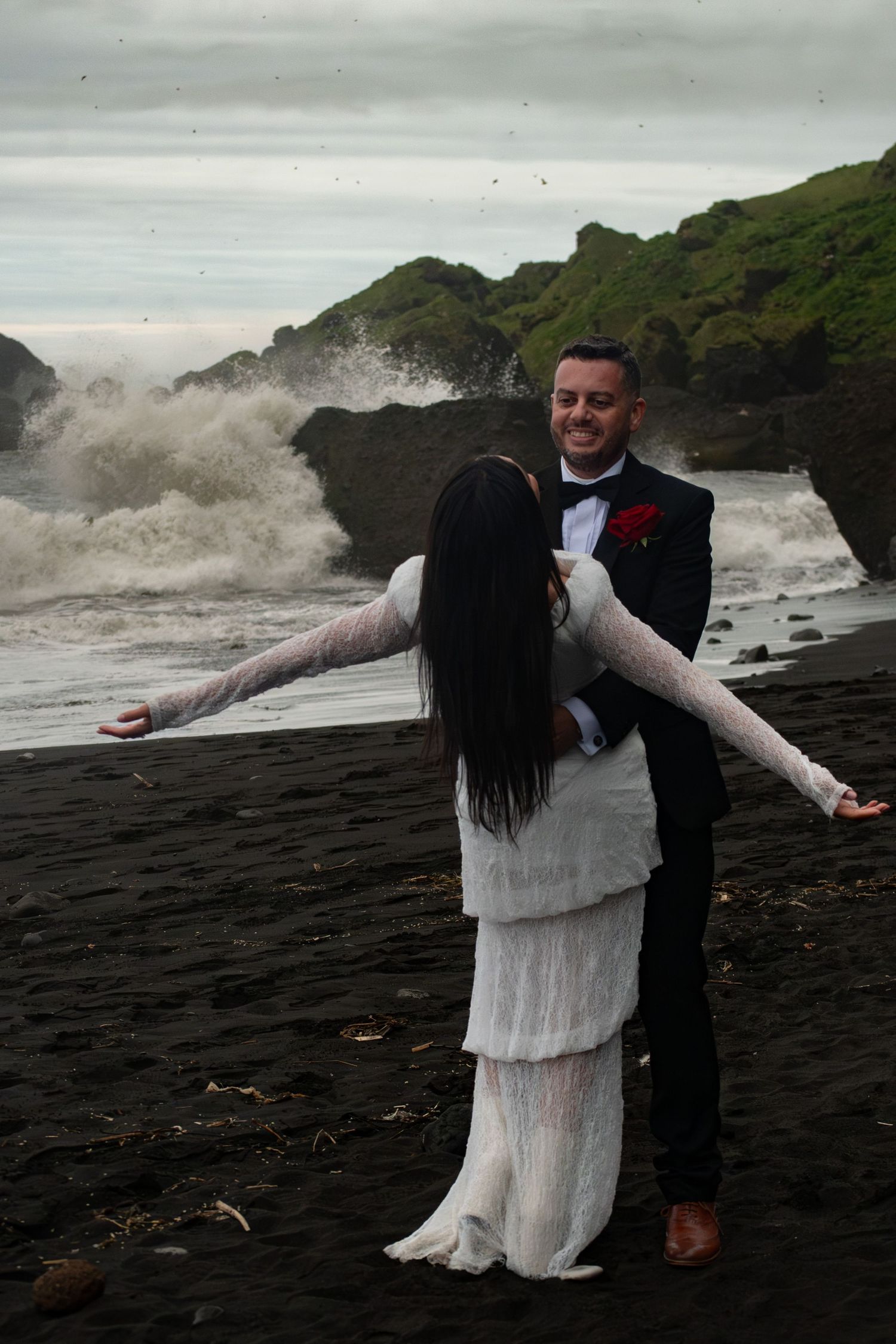 Bride and groom embrace on a black sand beach with dramatic crashing waves and green cliffs behind them.