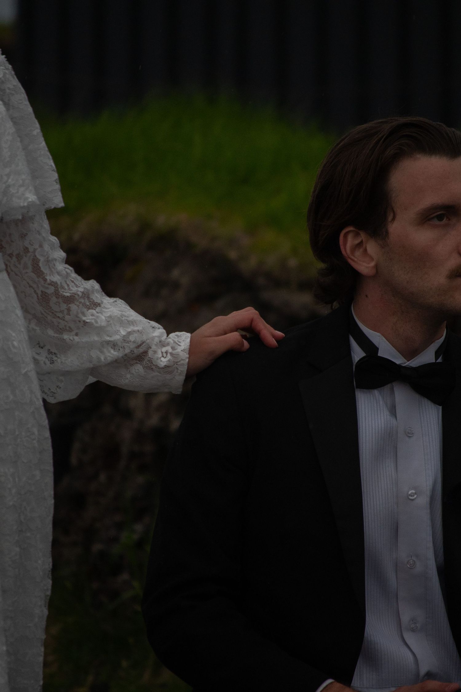 Bride in white lace dress placing hand on groom's shoulder in black tuxedo with bow tie outdoors.