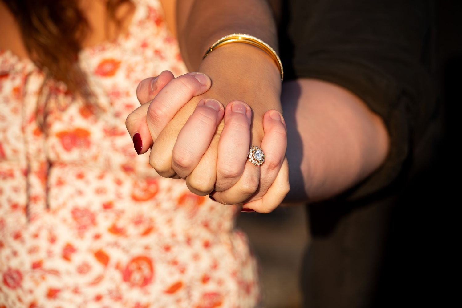 Close-up of couple holding hands showing engagement ring with diamond solitaire at golden hour.