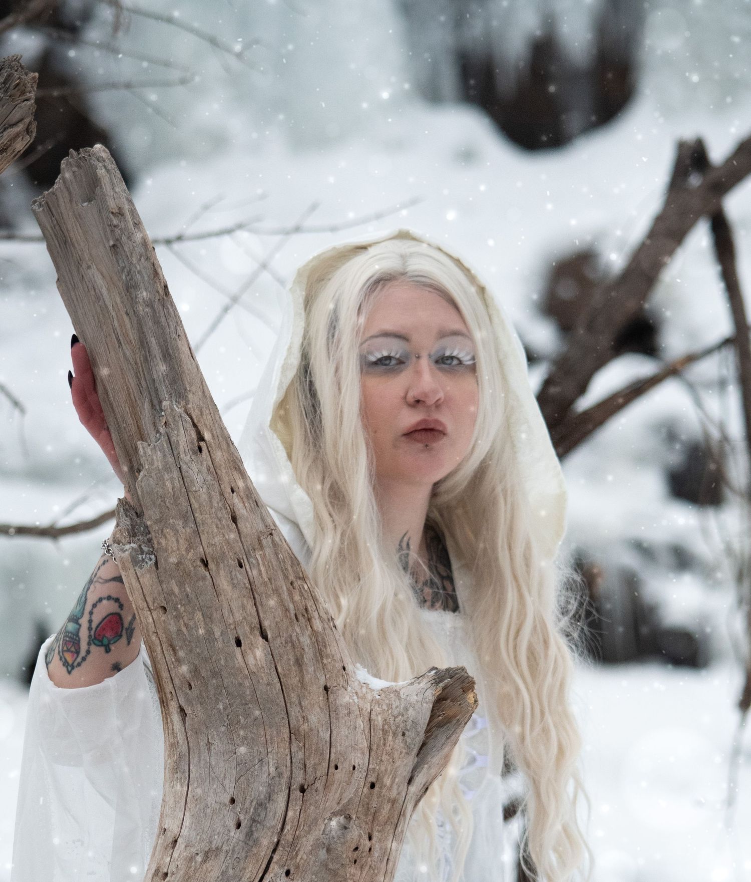 Tattooed blonde woman in white dress with silver eye makeup posing by driftwood in snow.