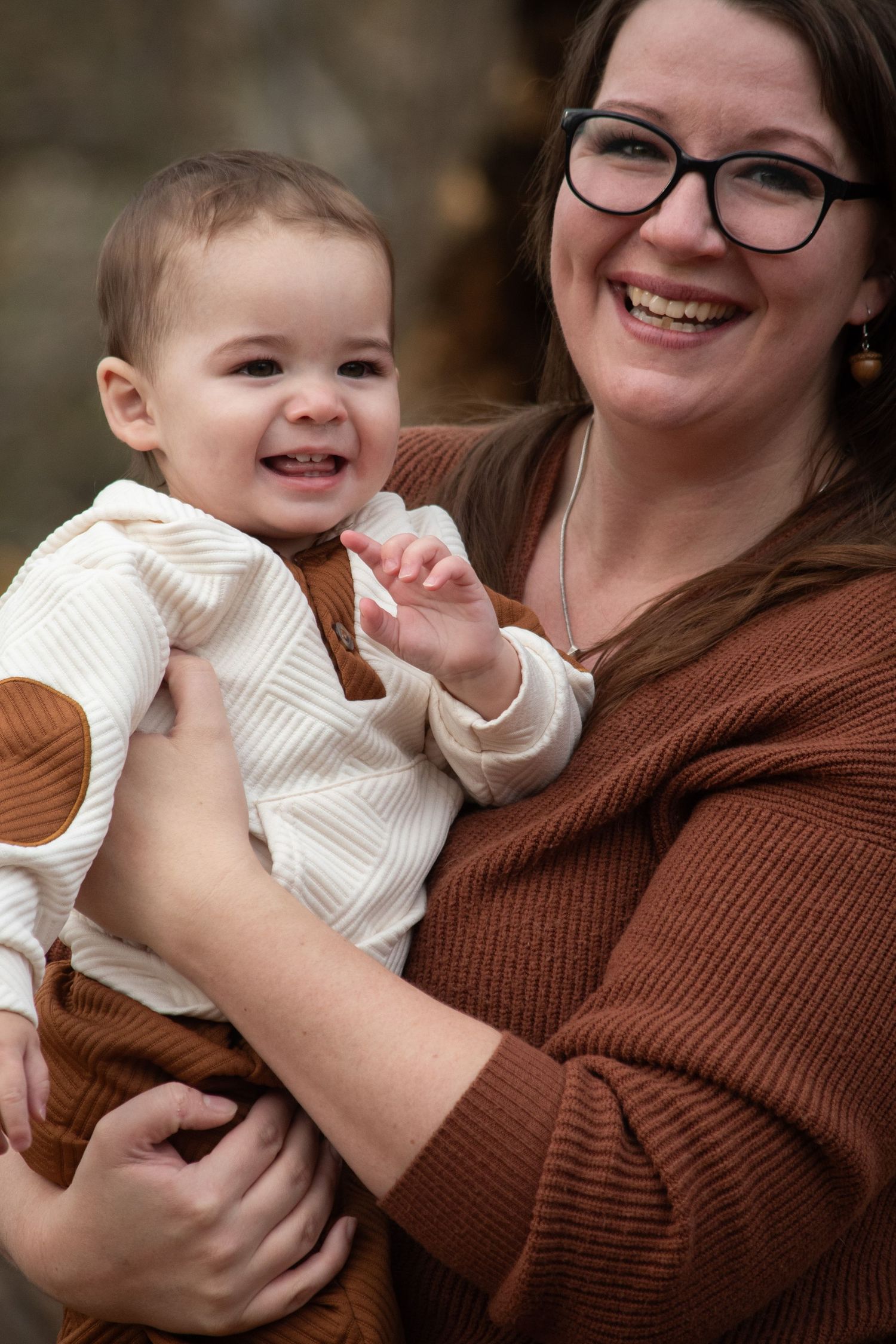 Smiling mother with glasses holding happy baby in cream striped outfit outdoors.