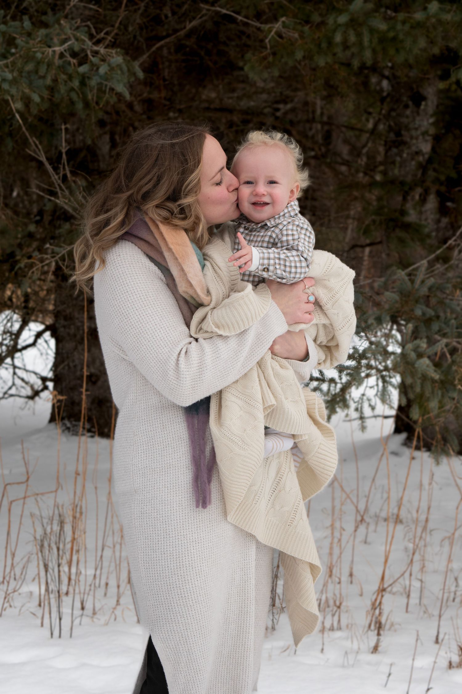 Mother in cream sweater kissing happy baby boy outdoors in snowy winter setting.