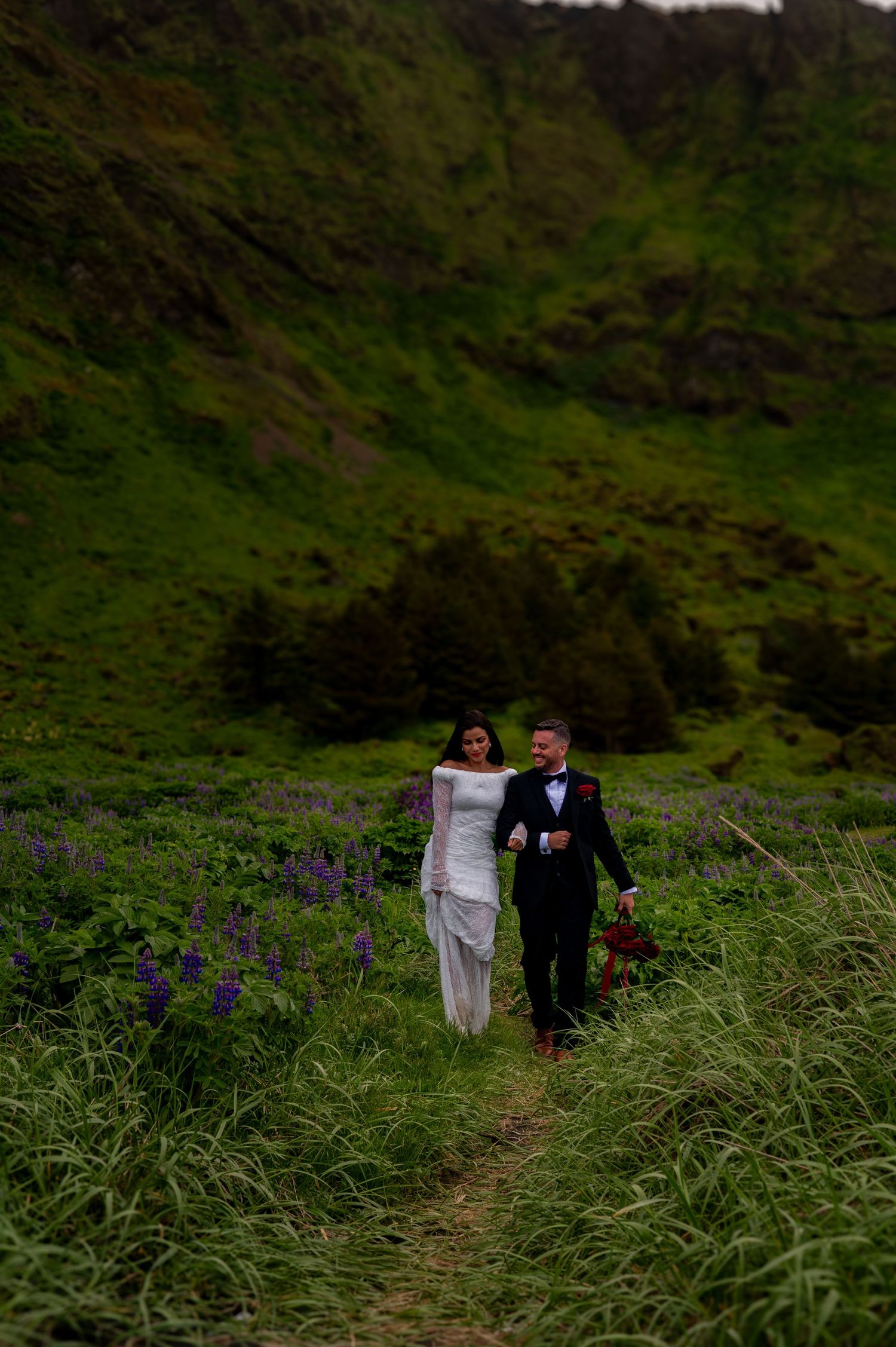Bride and groom walk through lush green meadow with purple wildflowers during Iceland elopement wedding.