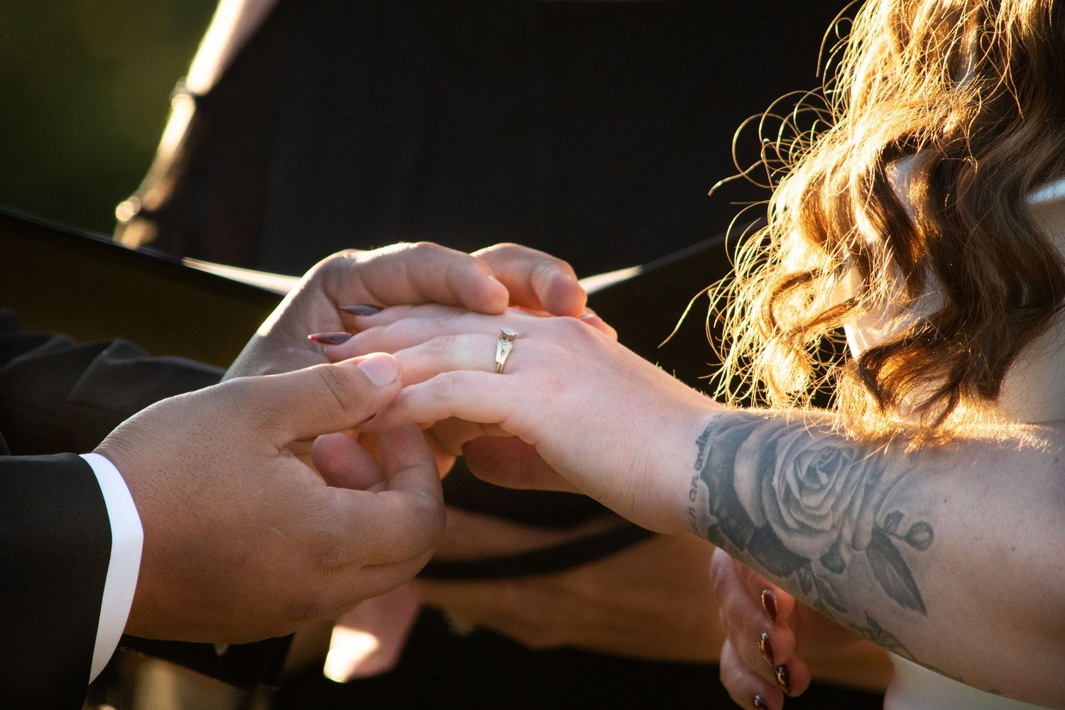 Groom sliding diamond ring onto bride's tattooed hand during wedding ceremony.