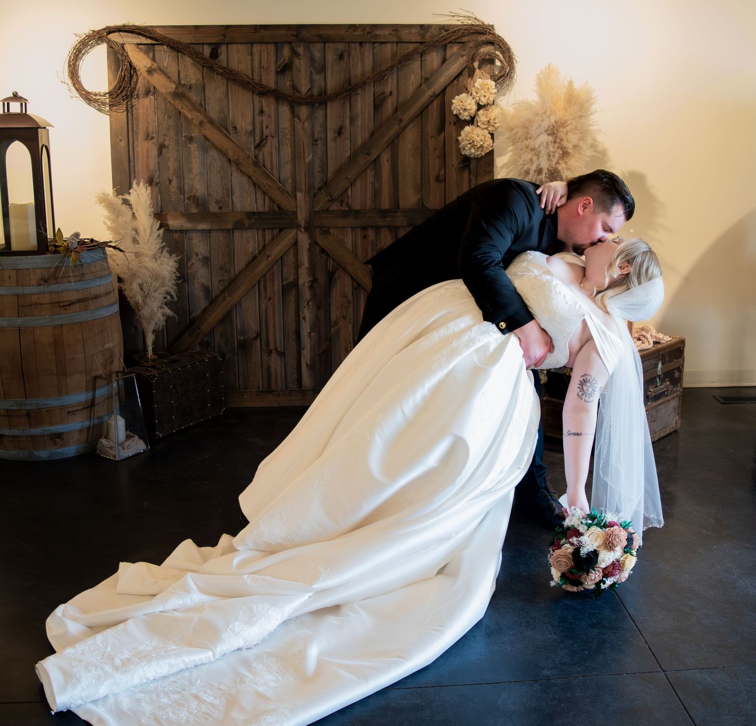 Groom dipping bride in elegant white gown for a romantic kiss in rustic barn wedding venue.