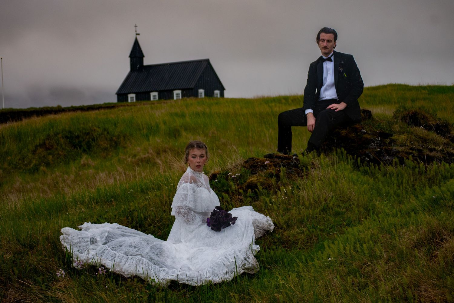 Bride in white gown and groom in suit pose on grassy hill with black church backdrop in Iceland.