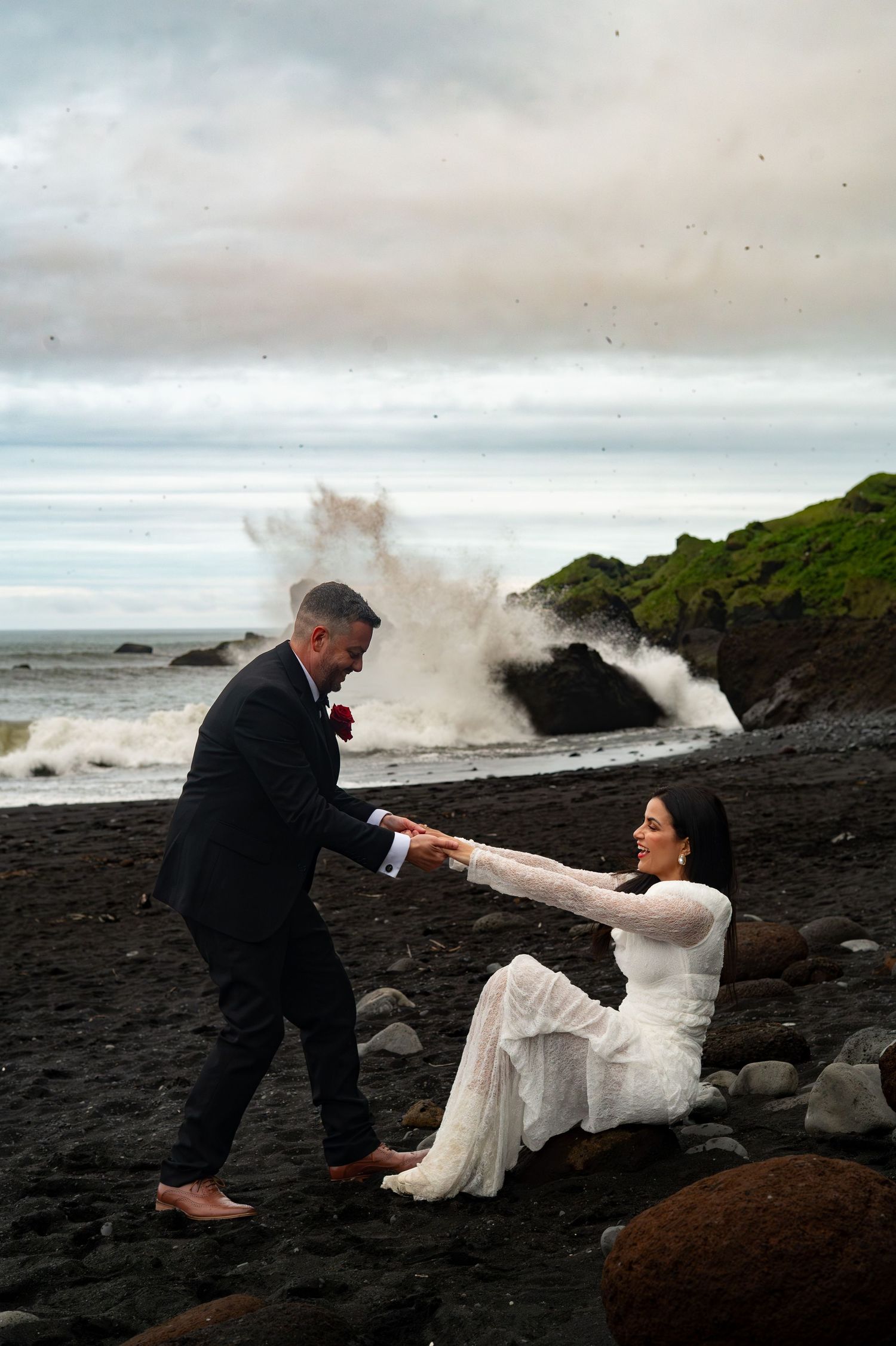 Groom helps bride rise from rocks on black sand beach as dramatic wave crashes behind them.