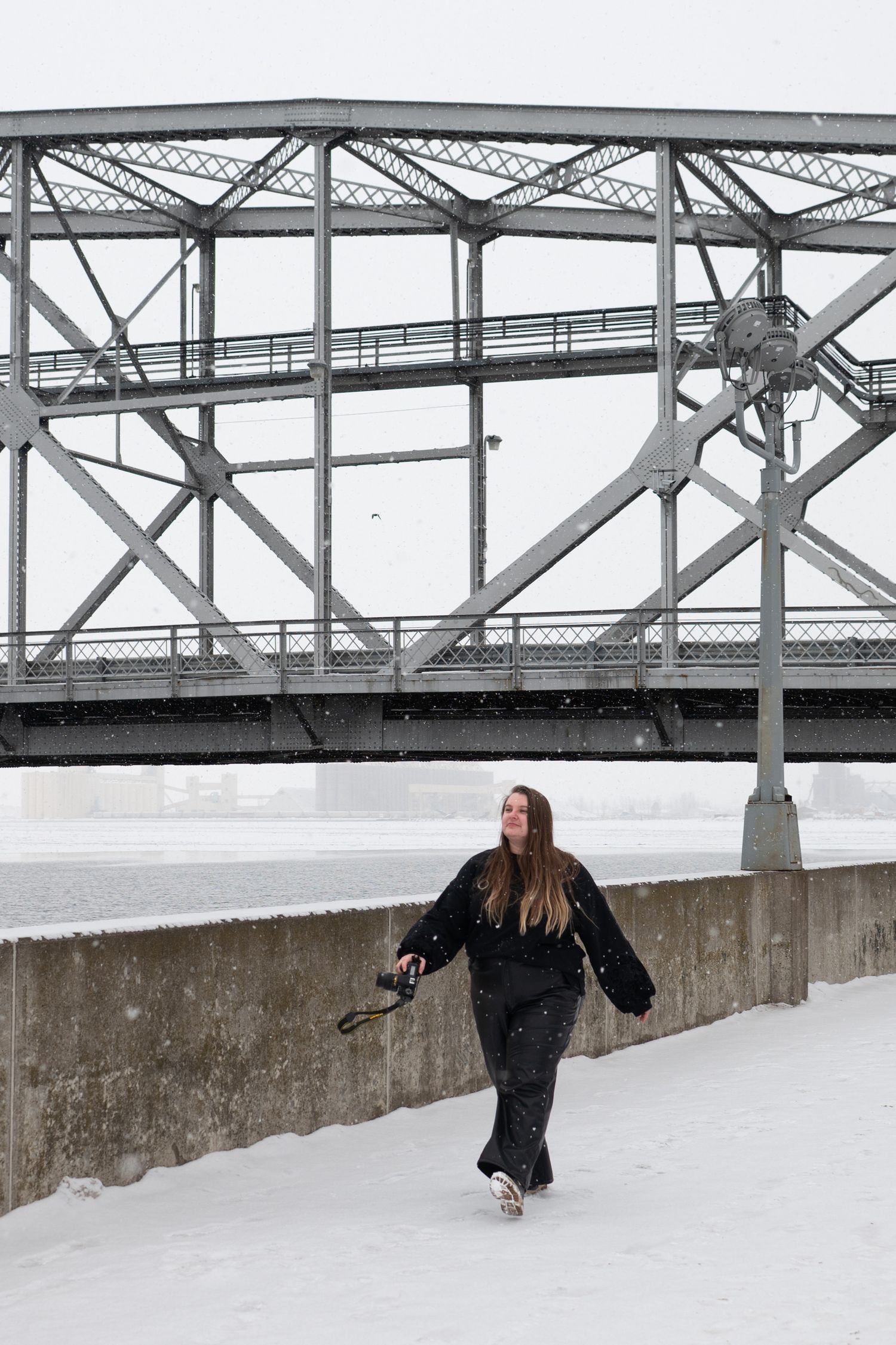 Woman in black outfit walking near snowy riverbank with steel bridge structure in background during winter snowfall.