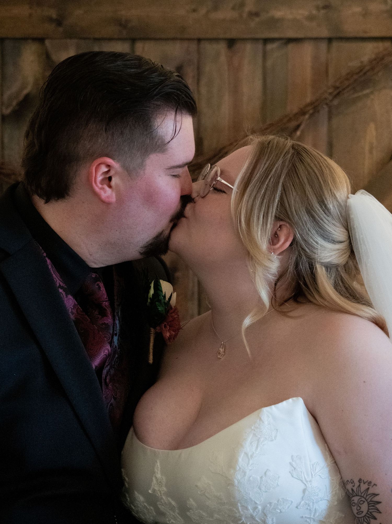 Newlyweds share romantic first kiss, bride in white lace gown and groom in black suit with burgundy tie.