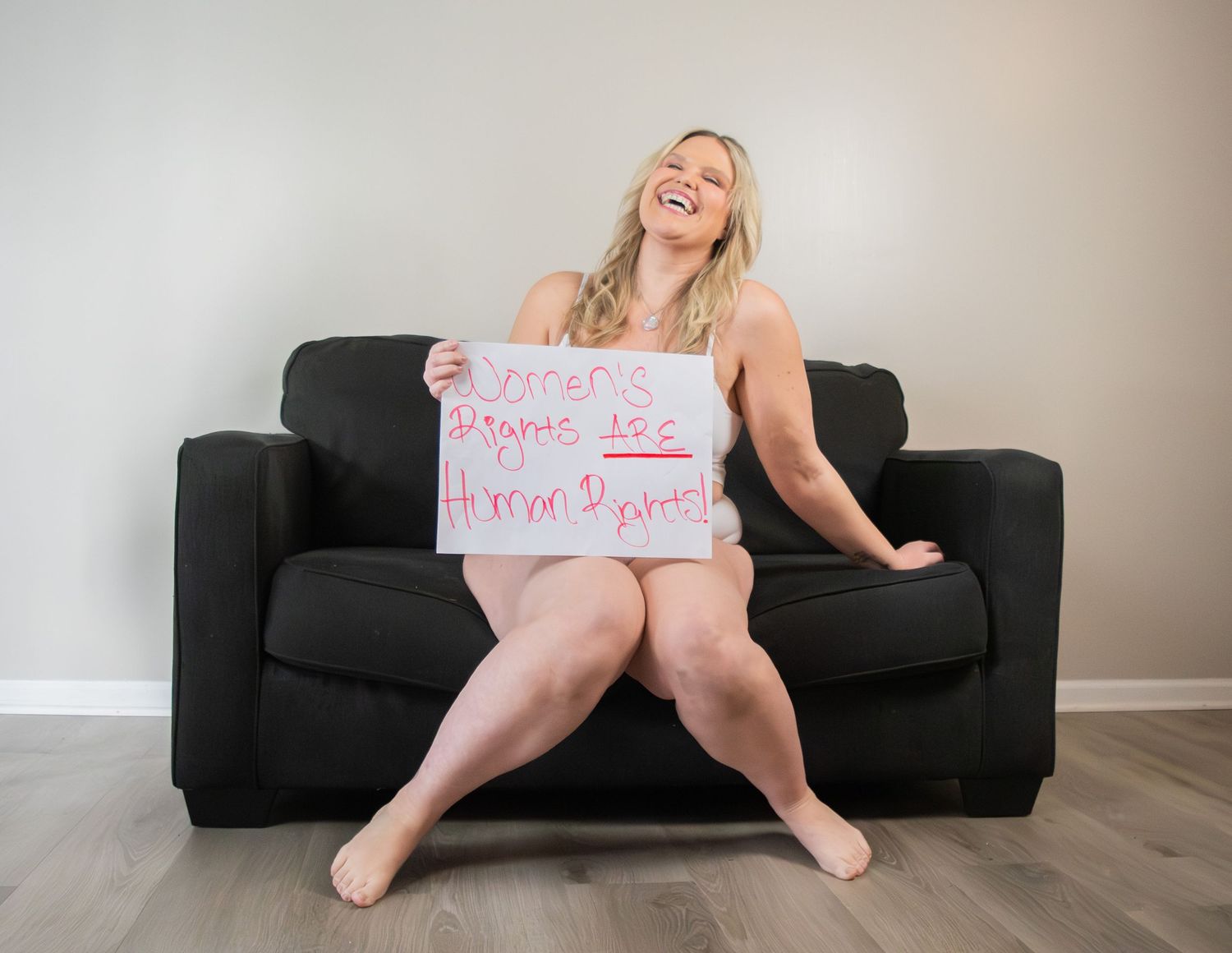 Laughing blonde woman holding a handwritten sign while sitting on a dark sofa.
