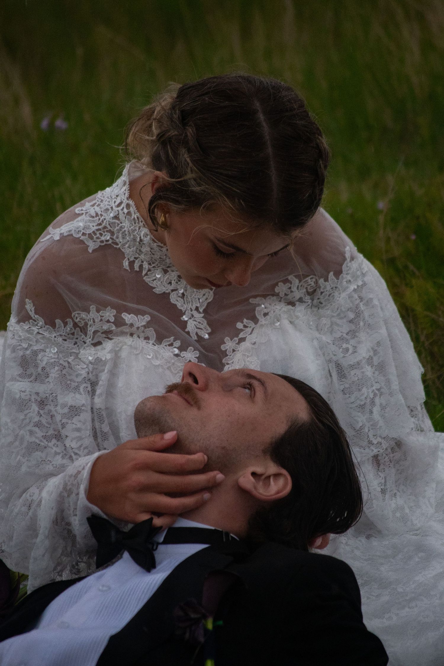 Bride in lace wedding dress tenderly cradling groom's face as he lies in green grass during romantic outdoor photo session.