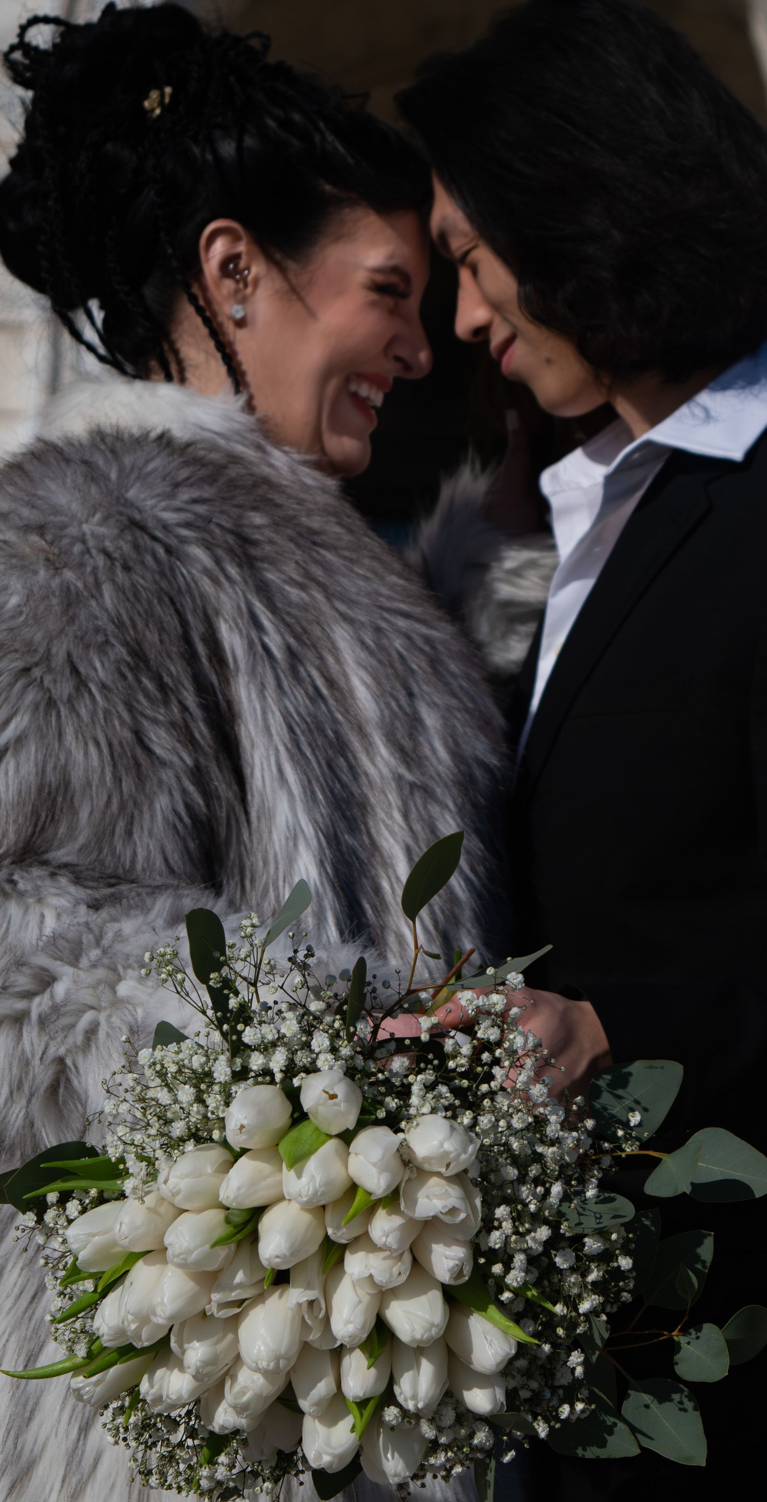 Two brides touch foreheads and smile joyfully, one holding white tulip bouquet with baby's breath.