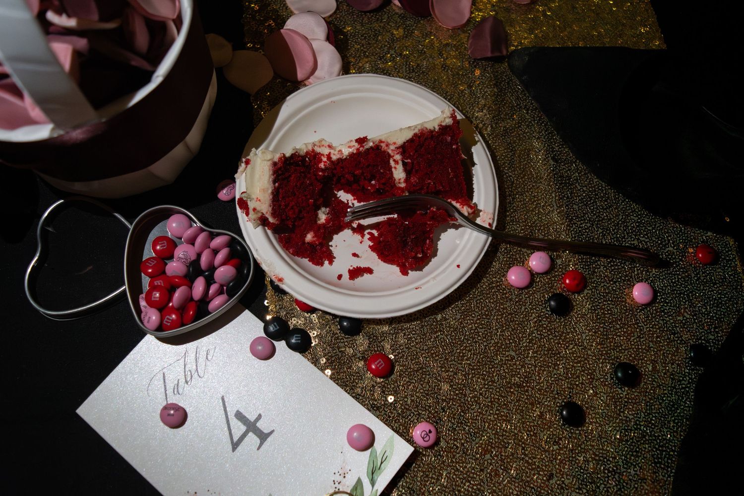 Slice of red velvet wedding cake on plate surrounded by colorful candies on glittery black table.