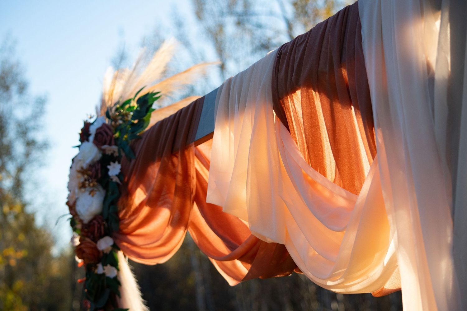 Elegant outdoor wedding arch with orange and white draped fabric and floral arrangement in sunlight.