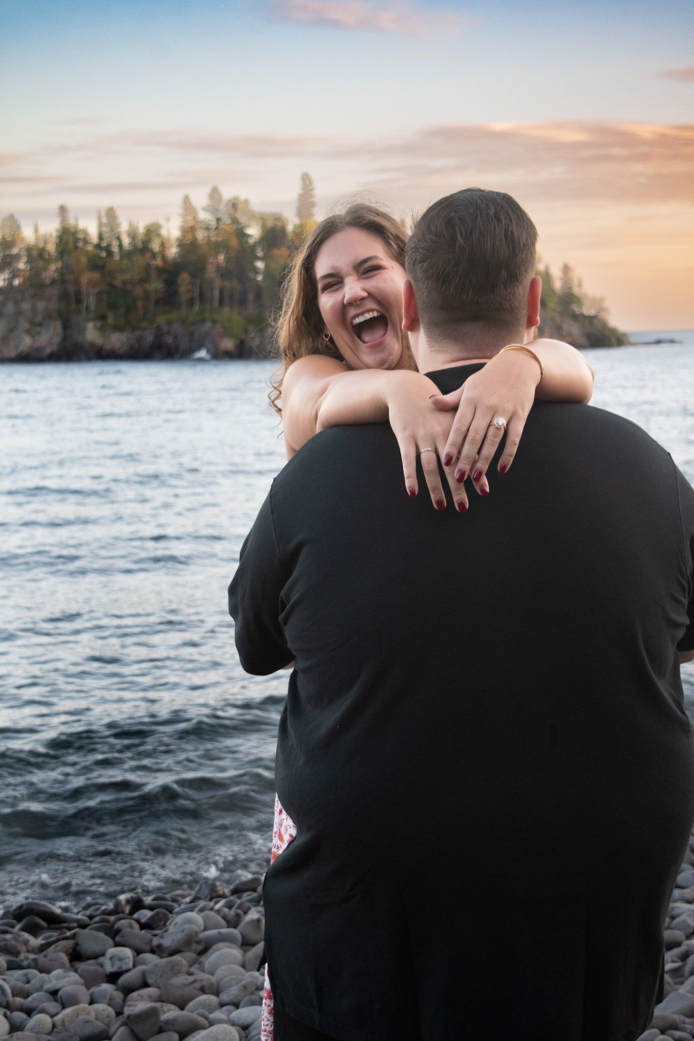 Newly engaged woman laughing with joy while embracing her partner on a pebble beach at sunset.