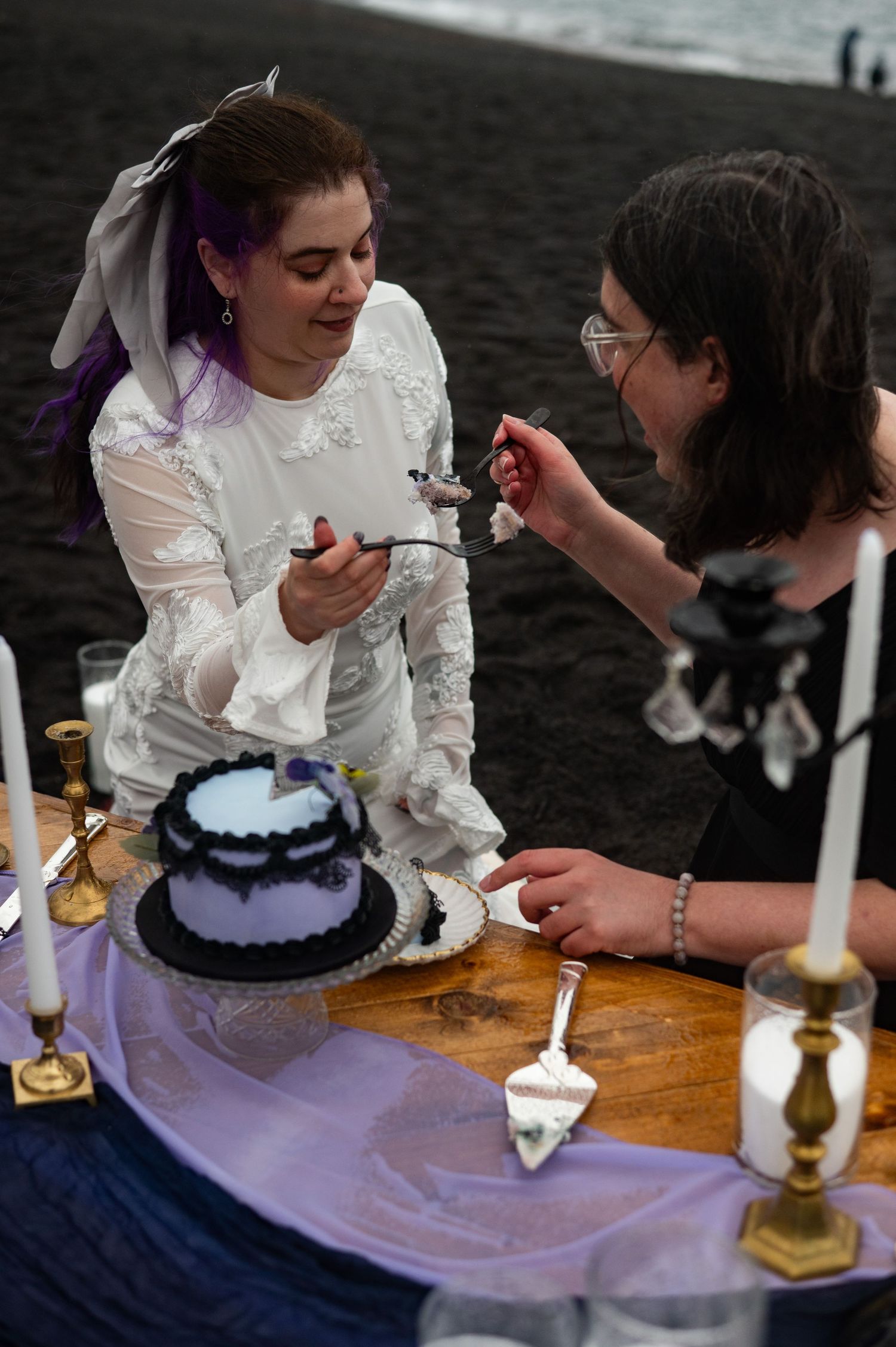 Bride in white dress feeds cake to guest at gothic black beach wedding reception with candles and dark cake.