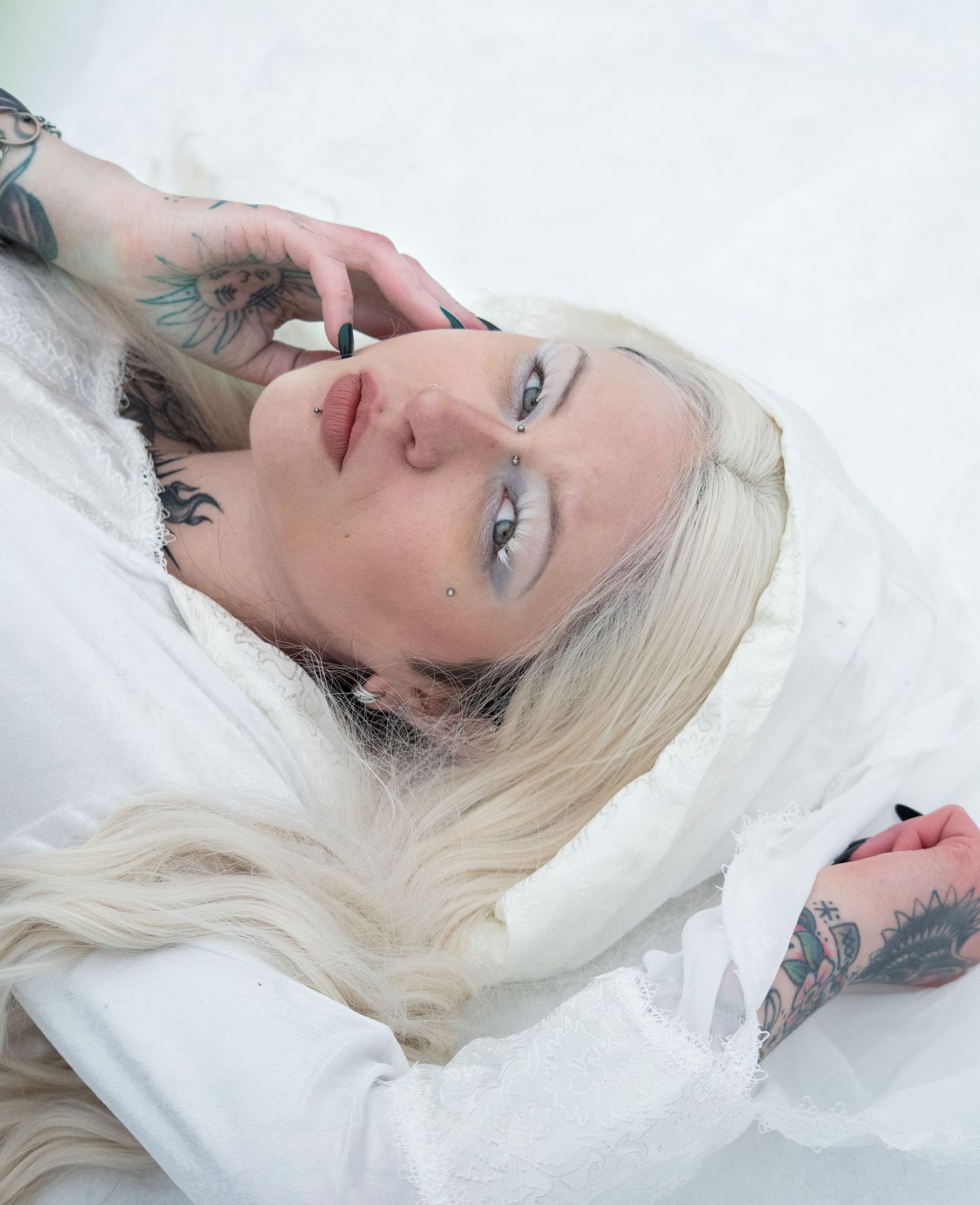 Tattooed blonde woman lying in snow wearing white dress with dramatic eye makeup.