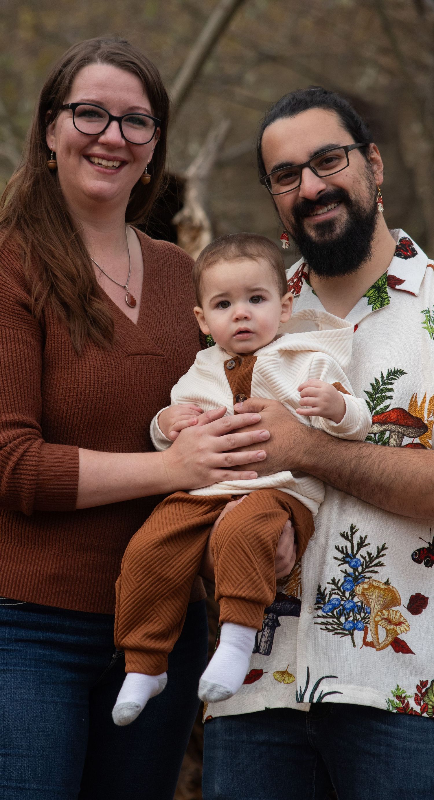 Smiling couple holding baby in cream outfit outdoors in autumn woodland.