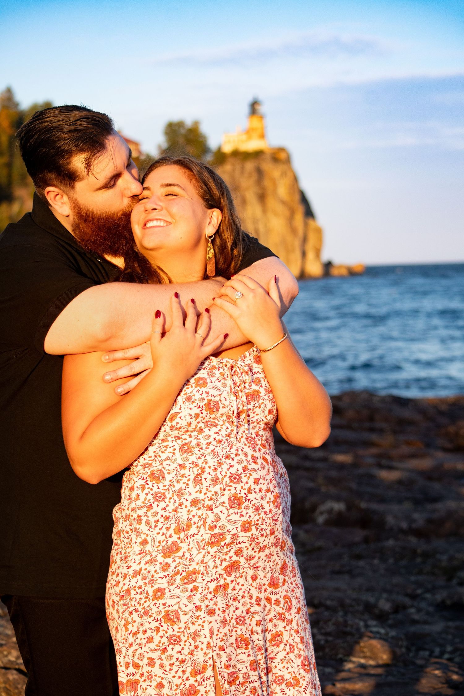 Couple embracing at sunset near Split Rock Lighthouse on Lake Superior, woman smiling joyfully.