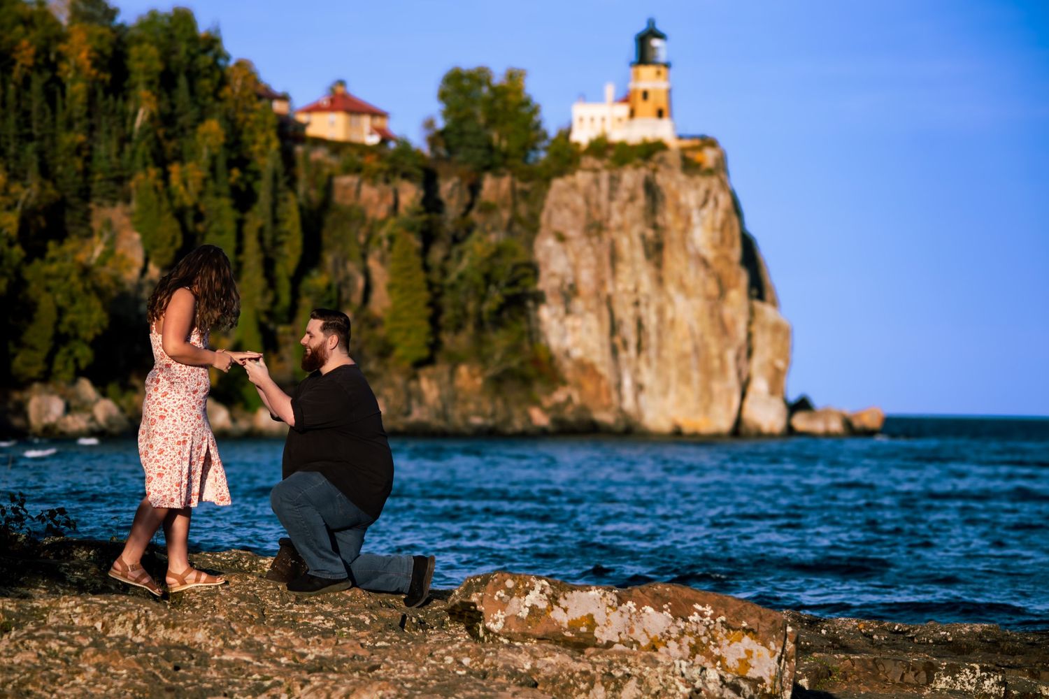 Man proposes to woman on rocky shore with Split Rock Lighthouse on cliff behind them.
