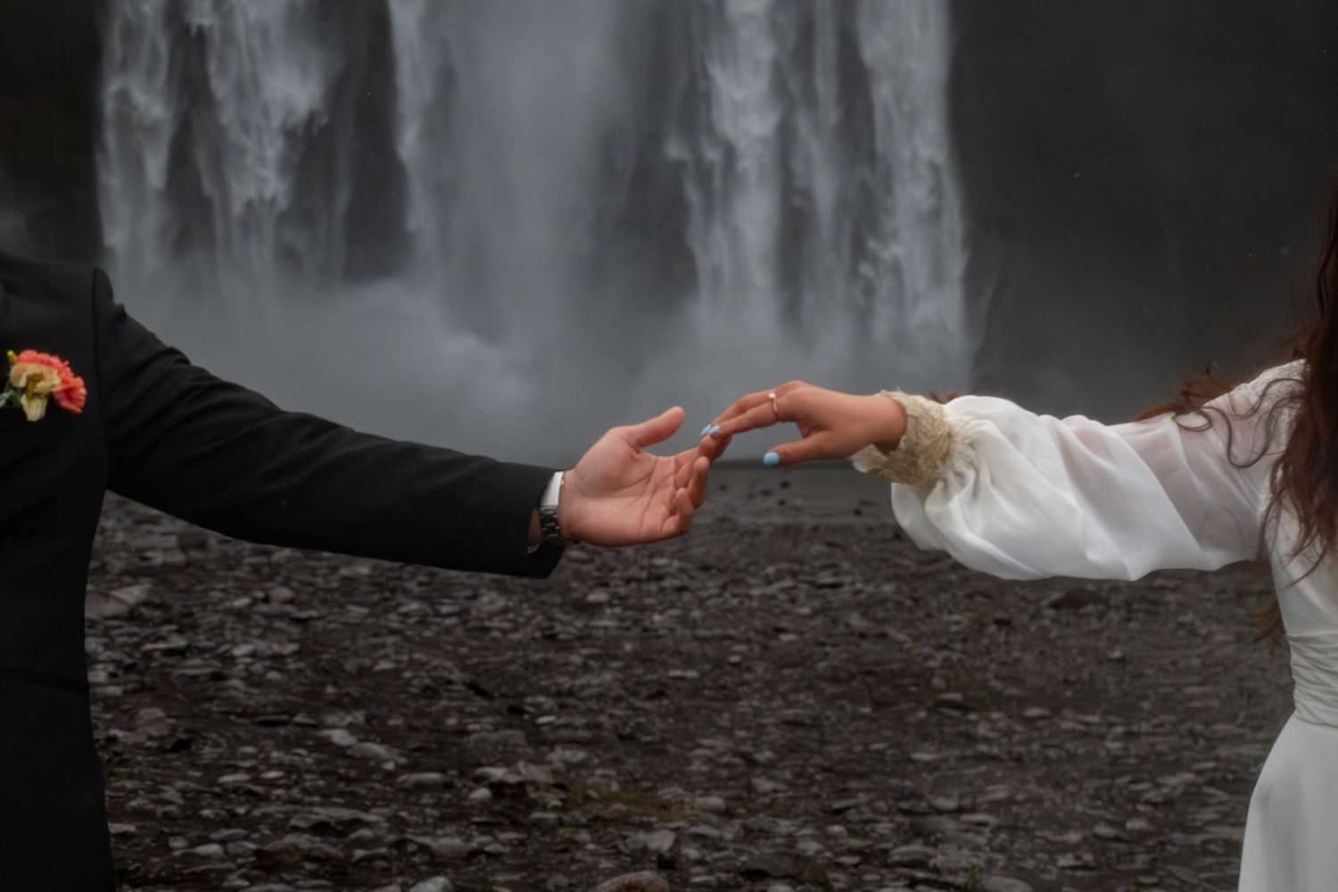 Couple reaching toward each other in front of a dramatic waterfall, bride in white lace dress and groom in black suit.