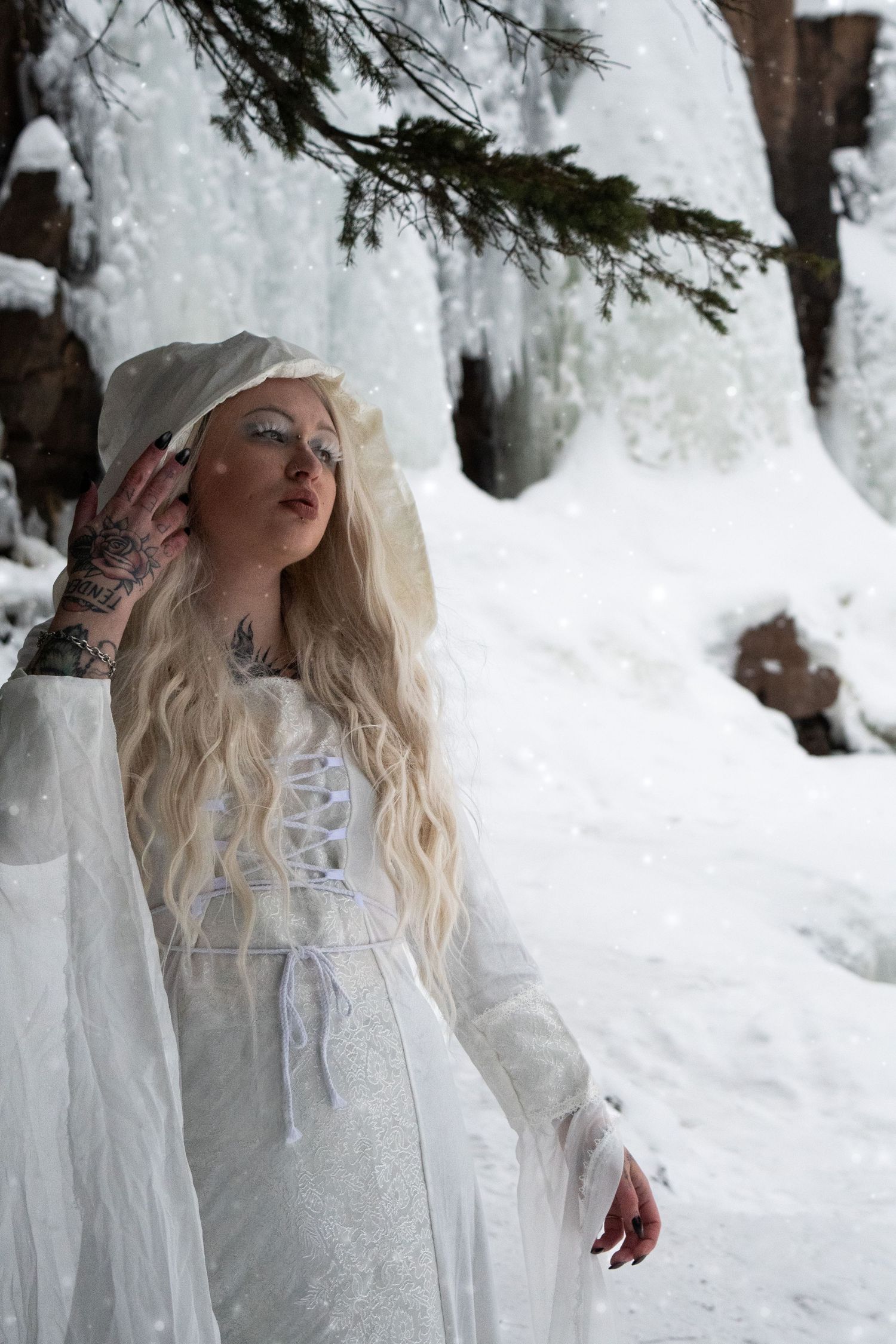 A blonde woman in a white lace gown and veil poses gracefully in a snowy winter forest with icy waterfalls.