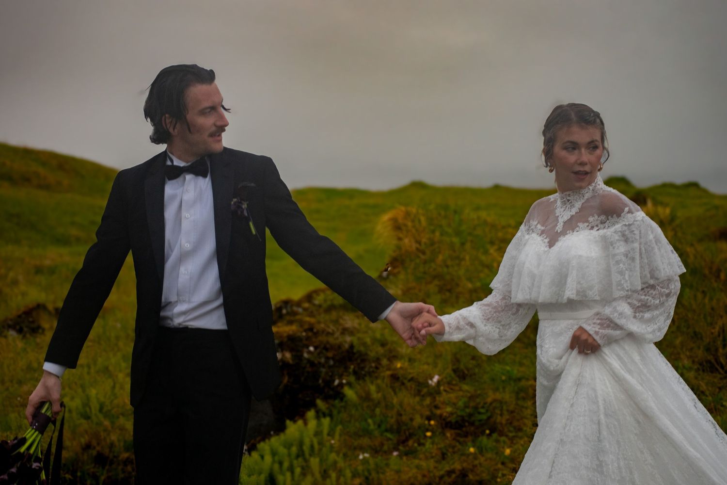 Bride in white lace gown holds hands with groom in black tuxedo on a lush green hillside.