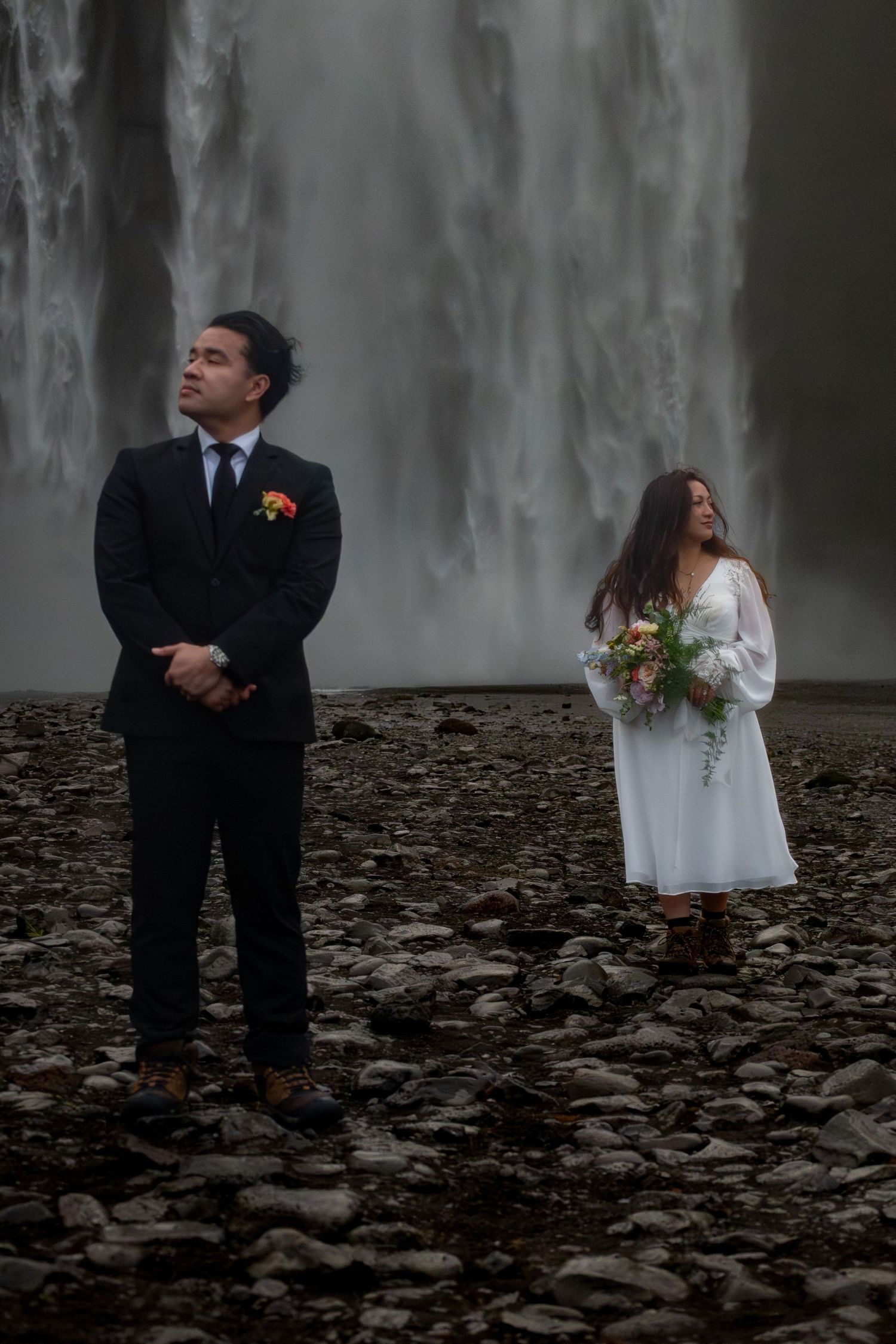 Couple in wedding attire standing before a majestic waterfall on rocky terrain, groom in black suit, bride in white dress.