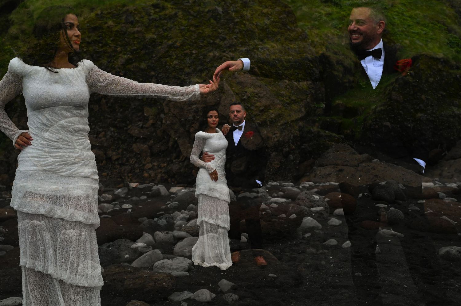 Bride in white lace gown and groom in tuxedo pose on rocky shore with green cliffs during outdoor wedding photoshoot.