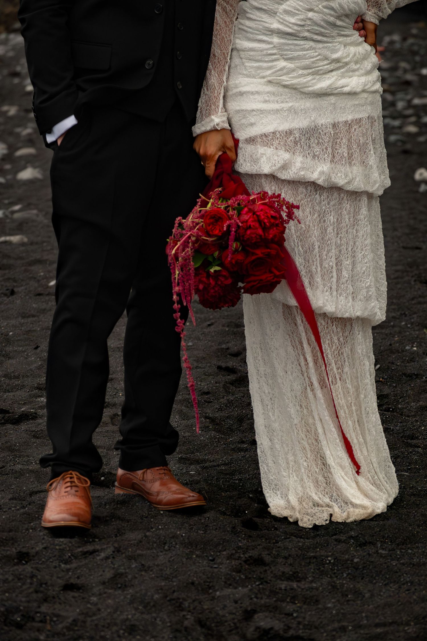 Bride in white lace gown holds red bouquet beside groom in black suit on dark sand beach.