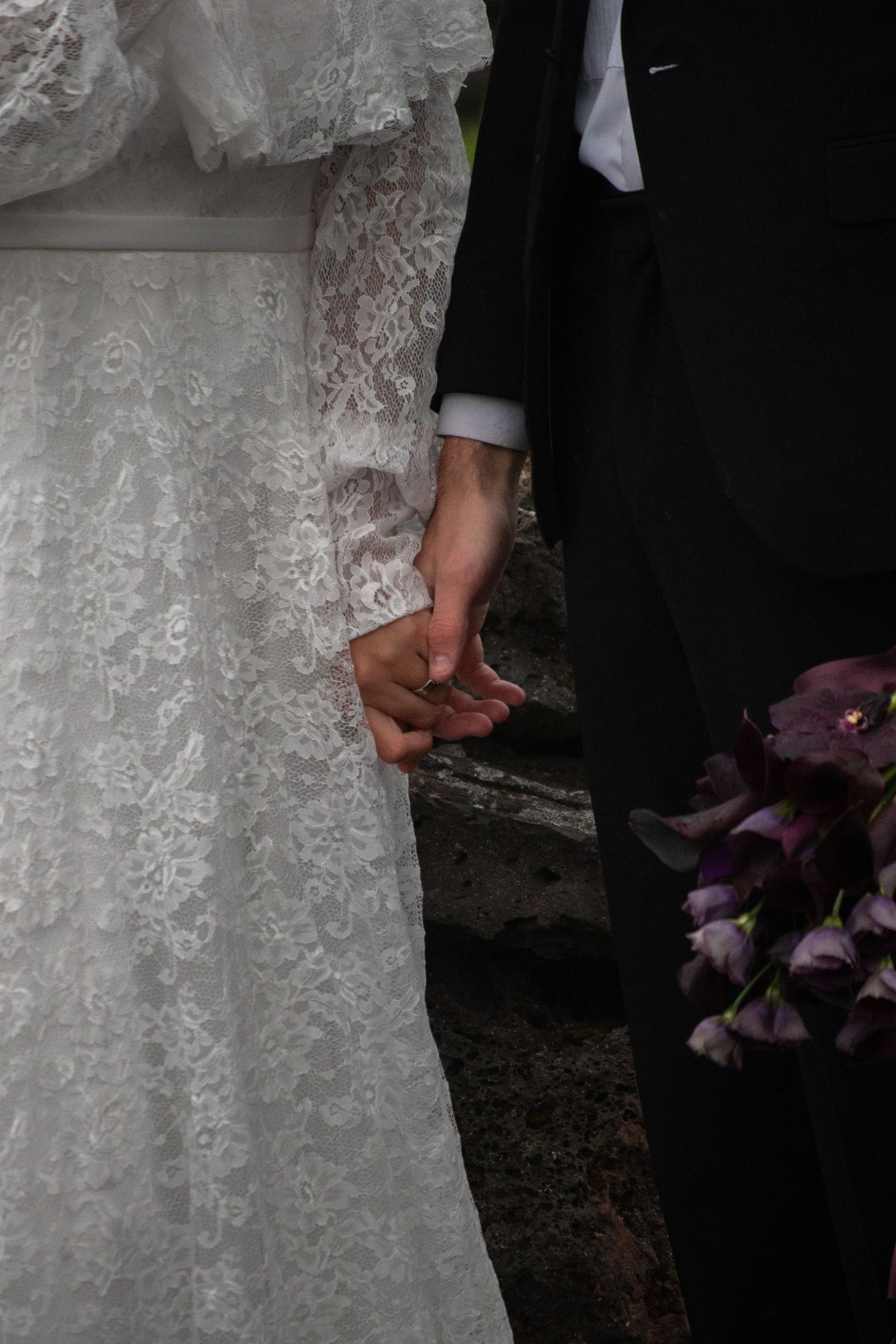 Bride and groom holding hands, white lace wedding dress and black suit, purple floral bouquet.