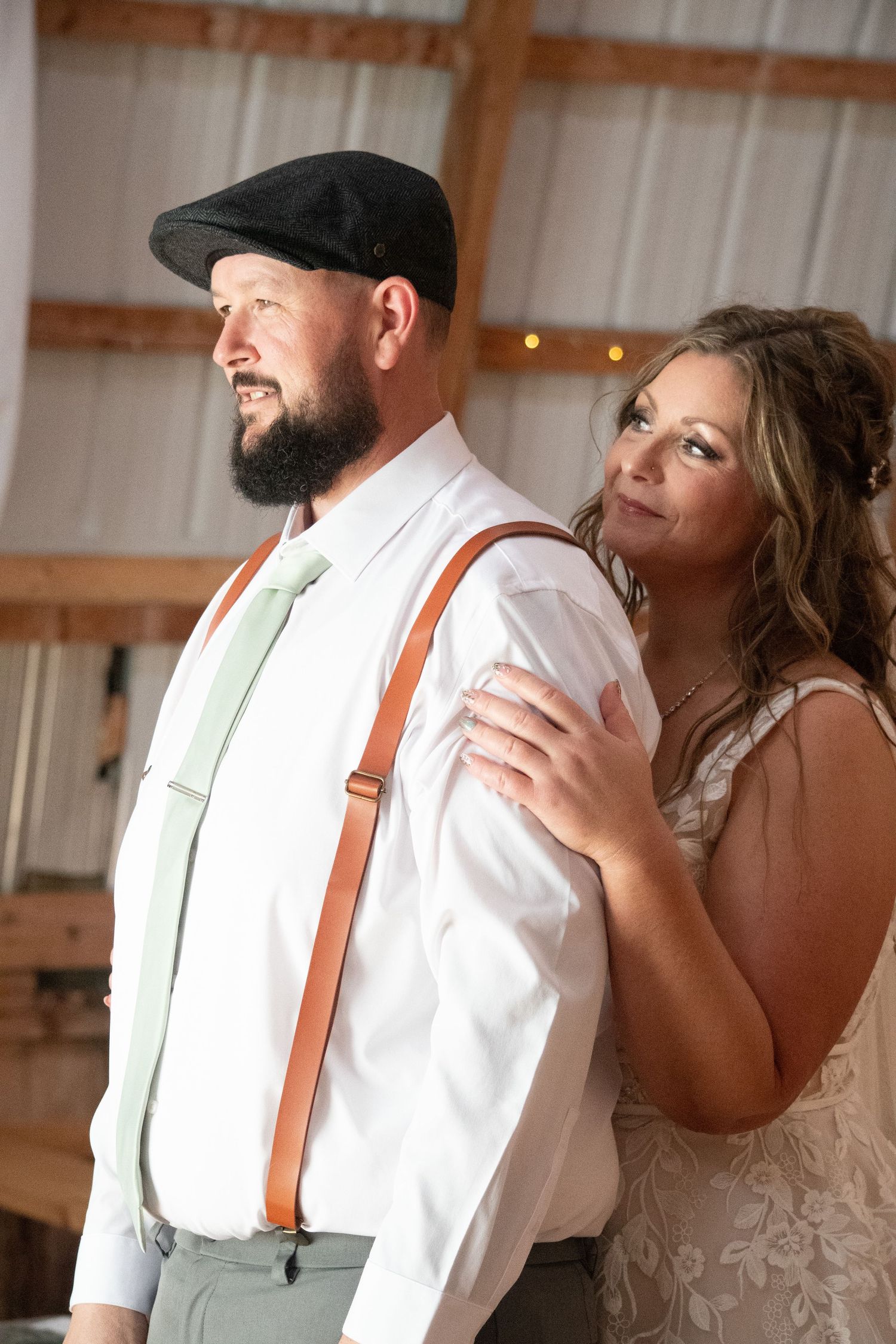 Bearded groom in flat cap with suspenders stands with bride leaning on his shoulder in barn venue.