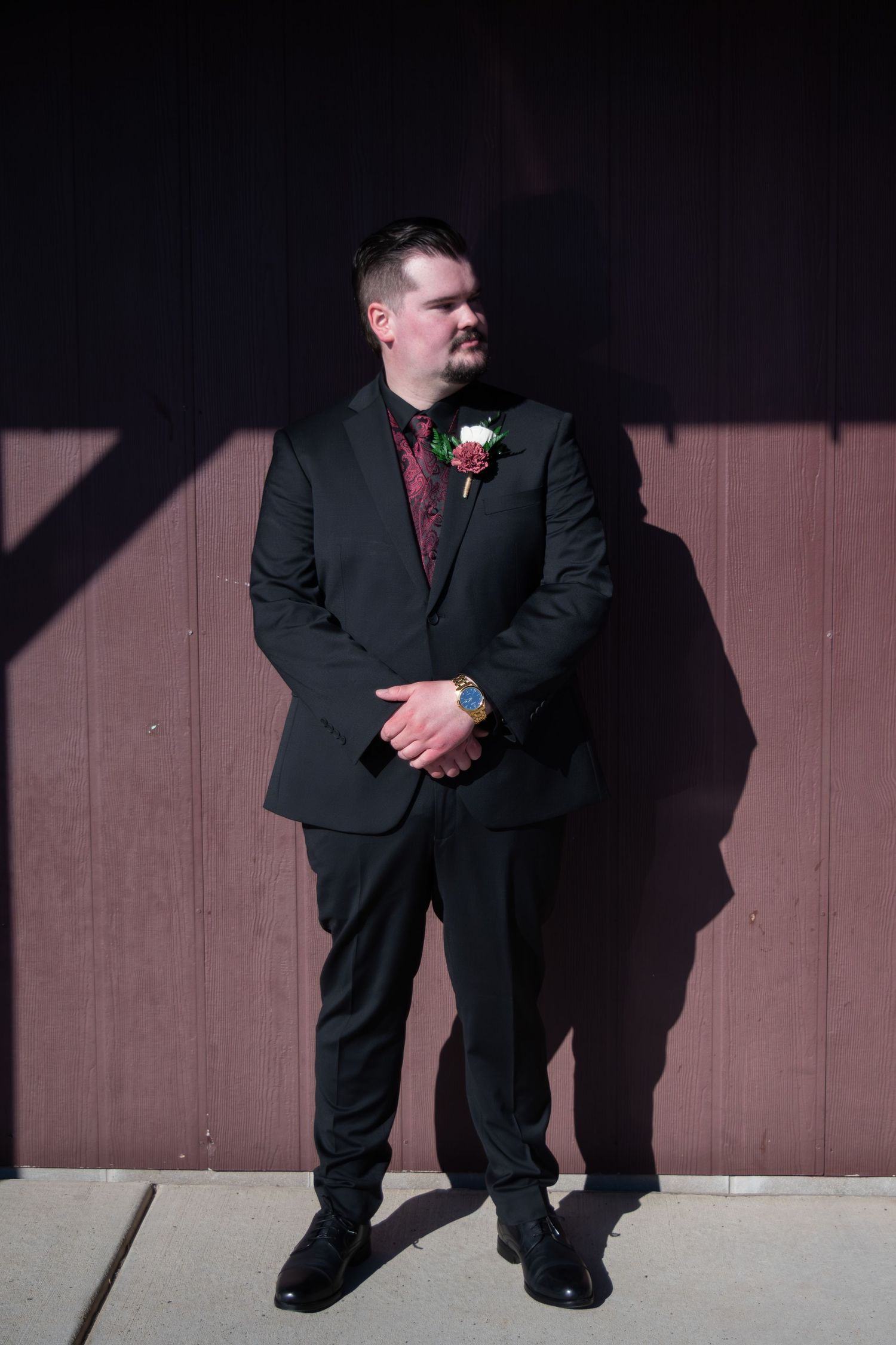 Dapper groom in black suit with burgundy paisley tie and floral boutonniere poses outdoors against wall.