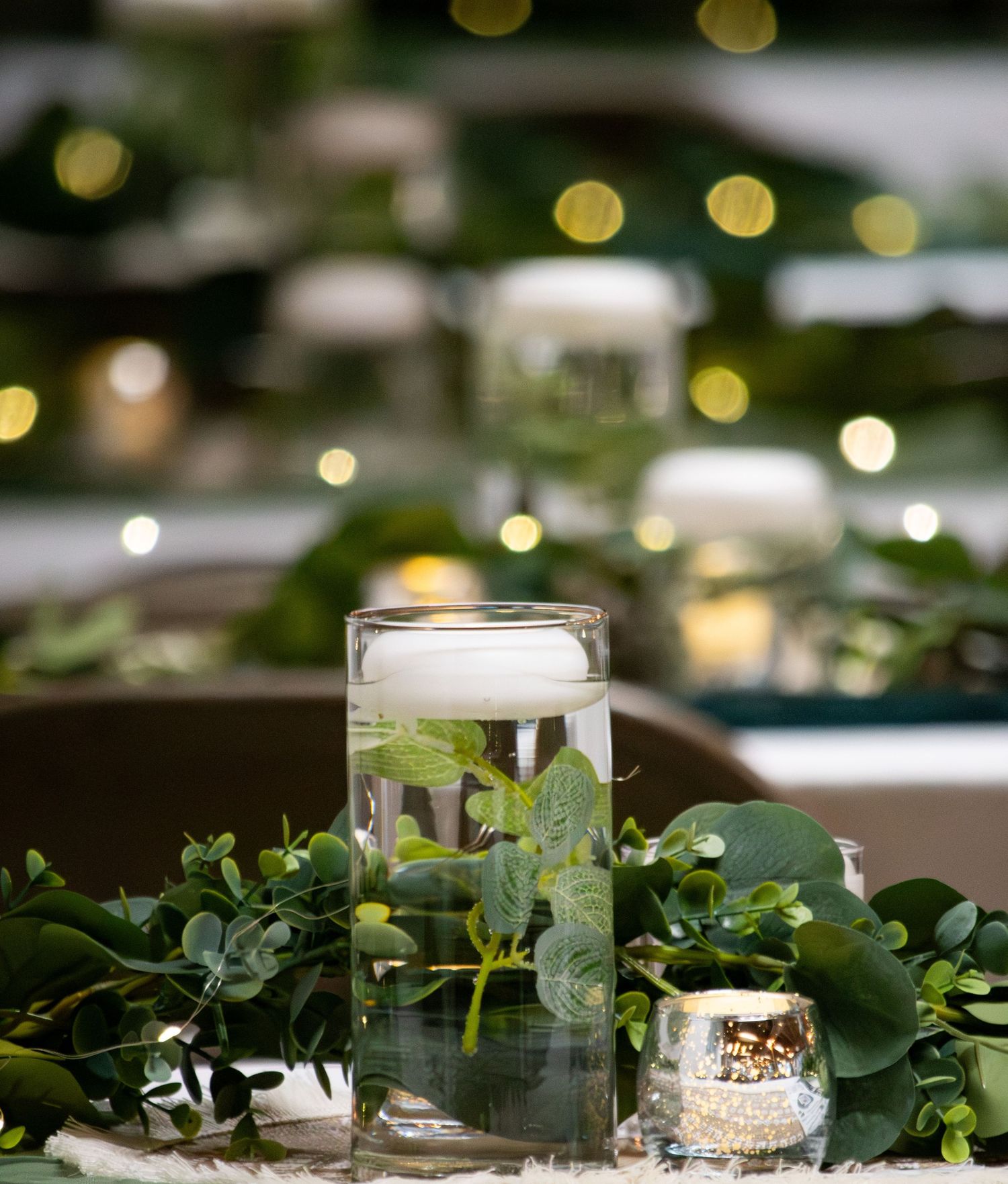 Elegant wedding centerpiece with floating candle in glass cylinder vase surrounded by lush green eucalyptus garland.