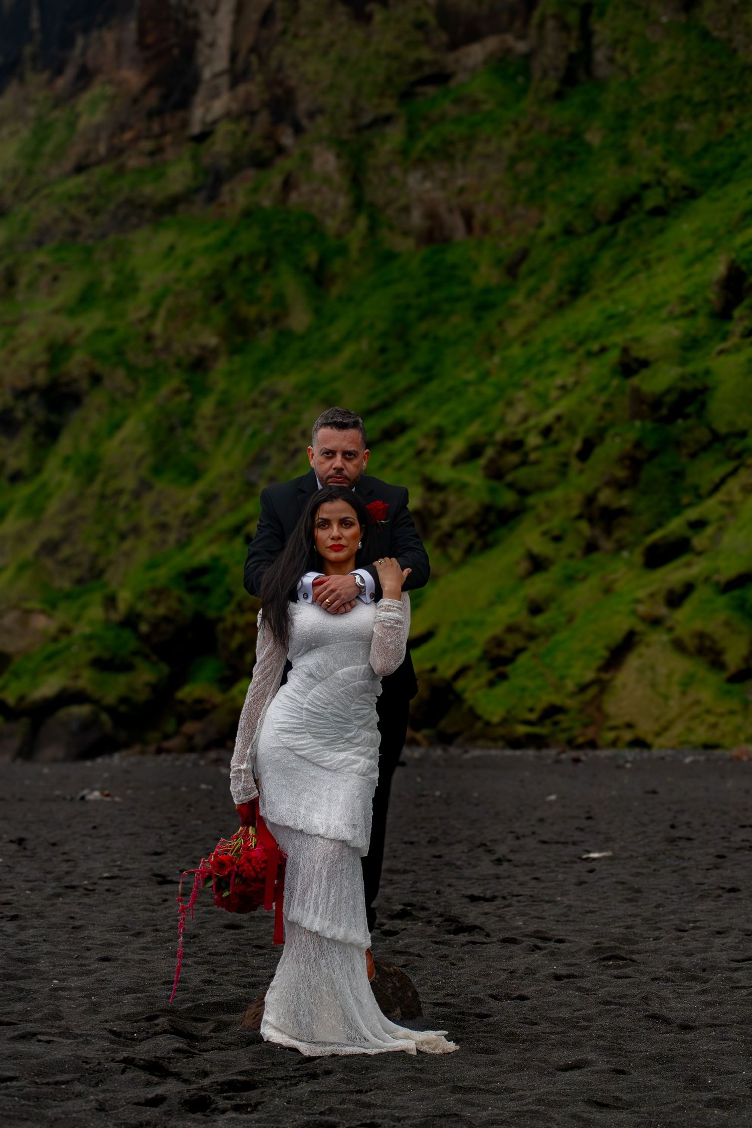 Wedding couple embraces on black sand beach with green cliffs, bride holds red bouquet.
