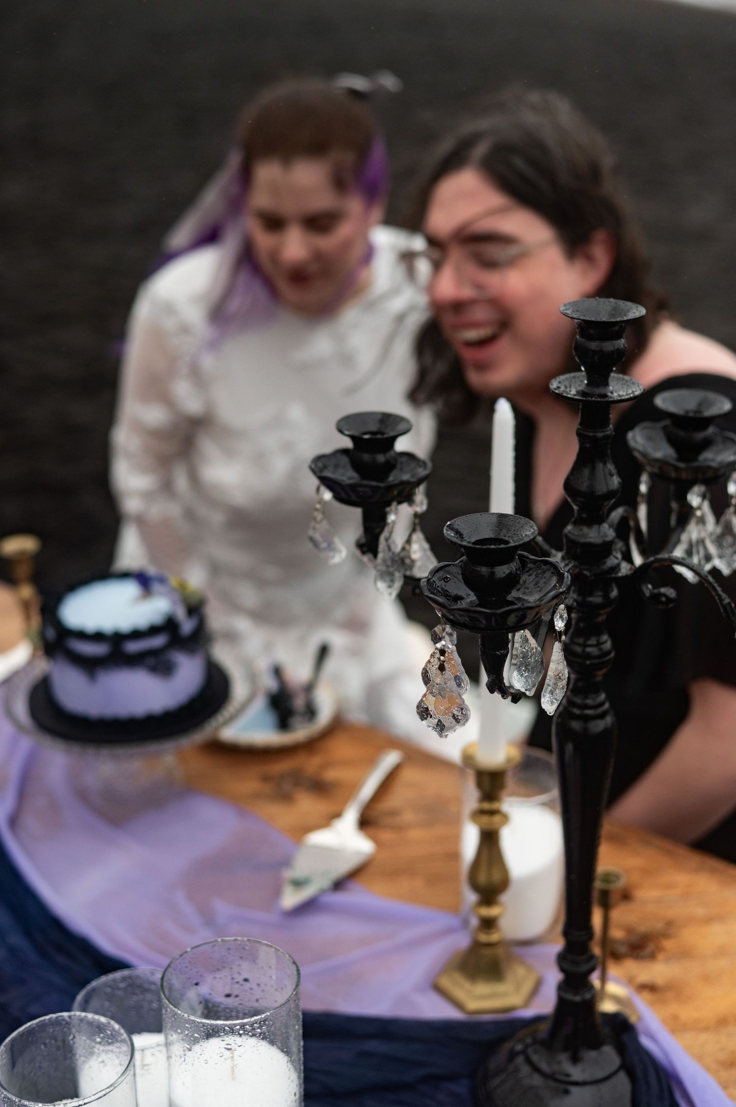 Two women celebrate at a gothic-themed wedding table with black candelabra and a dark blue cake.