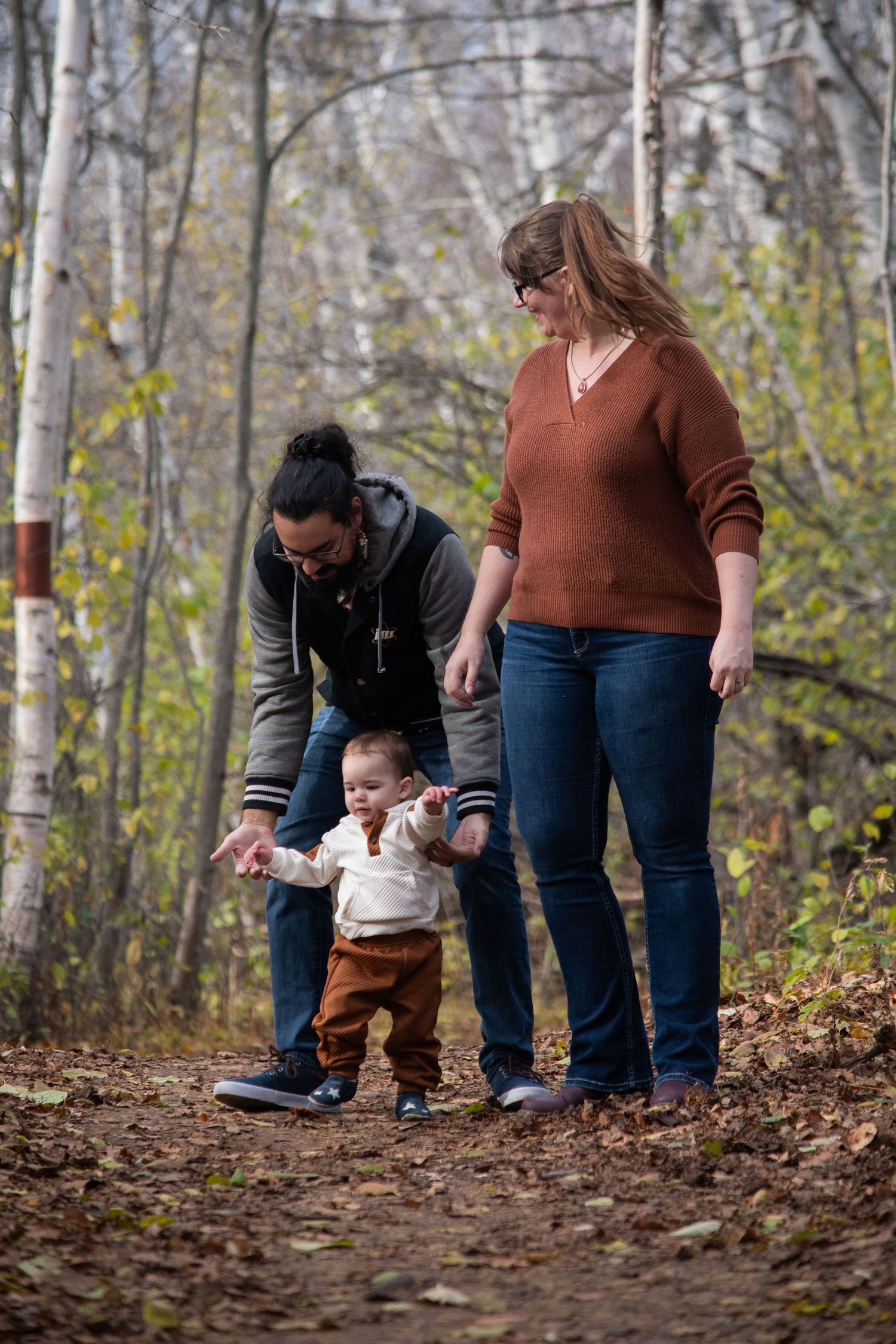 Family with toddler taking first steps on autumn forest trail, parents guiding child through fallen leaves.