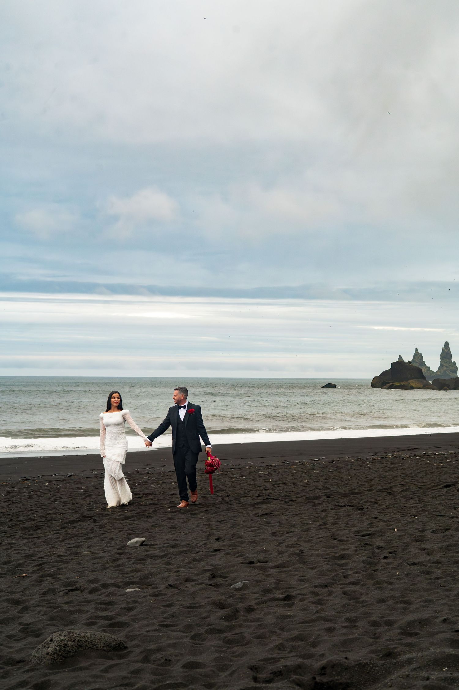 Bride and groom walk hand-in-hand on black sand beach with sea stacks in background.