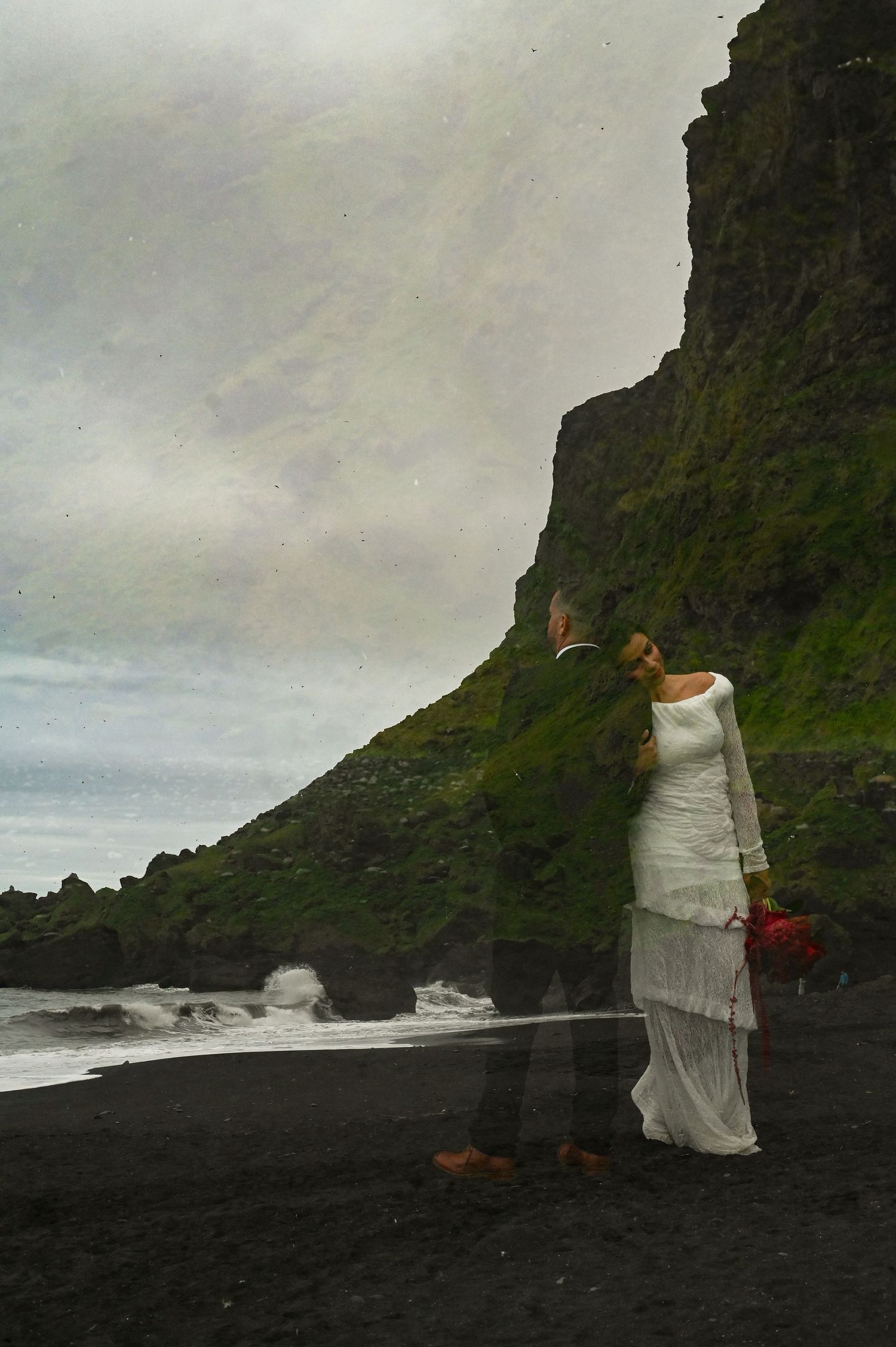 A bride and groom share a romantic embrace on a dramatic black sand beach with a mossy cliff backdrop.