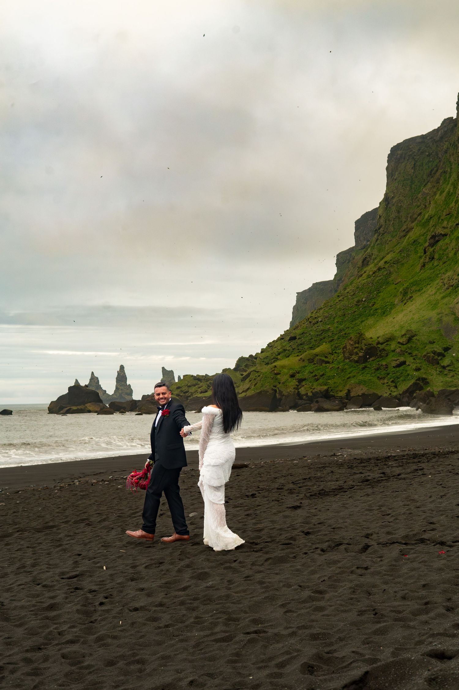 Couple in wedding attire on Iceland's black sand beach, with dramatic cliffs and sea stacks in the background.